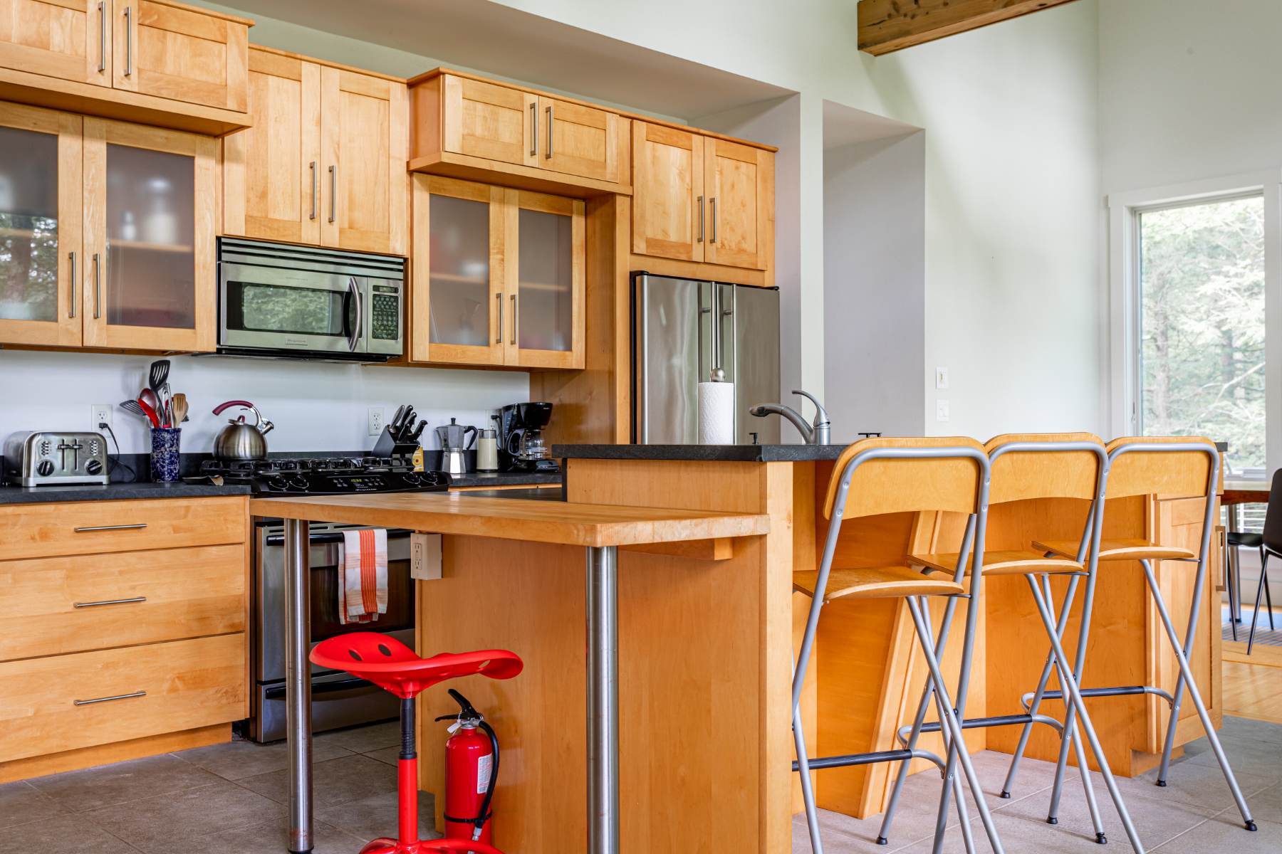 A bright kitchen features light wood cabinetry, black countertops, and a large island with bar seating and wooden stools. A red fire extinguisher is visible near the island, and stainless steel appliances are integrated into the functional and open layout.