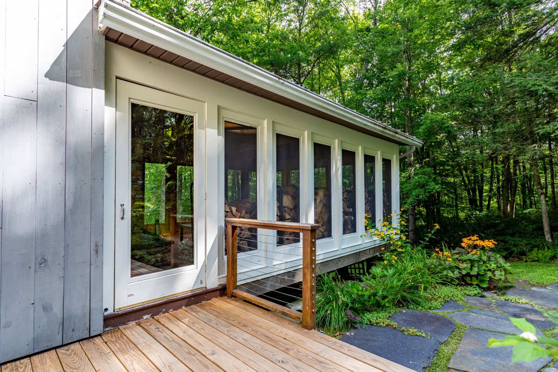 A wooden deck with a simple railing leads to a screened-in porch area, offering views of the surrounding lush green trees, with stacked firewood visible through the screens.