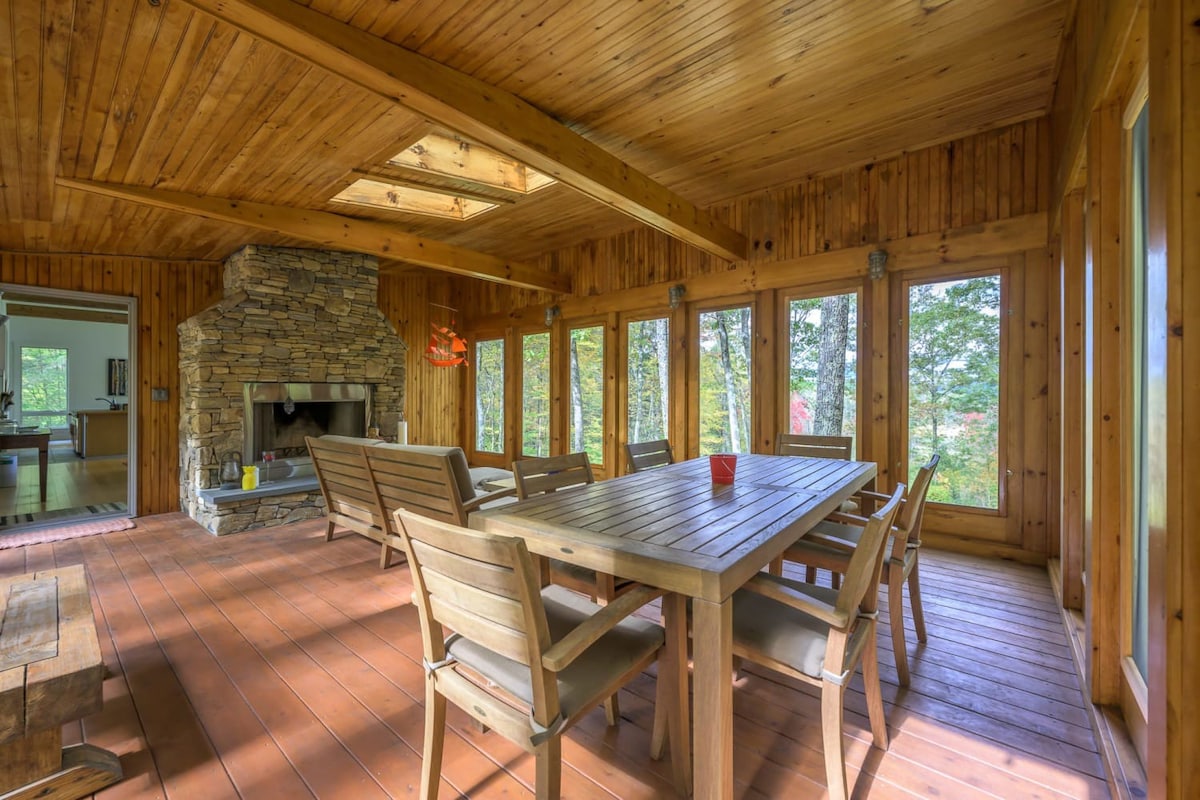 A rustic dining area in a sunroom features a large wooden dining table surrounded by matching chairs, set against a prominent stone fireplace. The room boasts extensive wooden paneling on walls and ceiling, with large windows overlooking a dense forest.