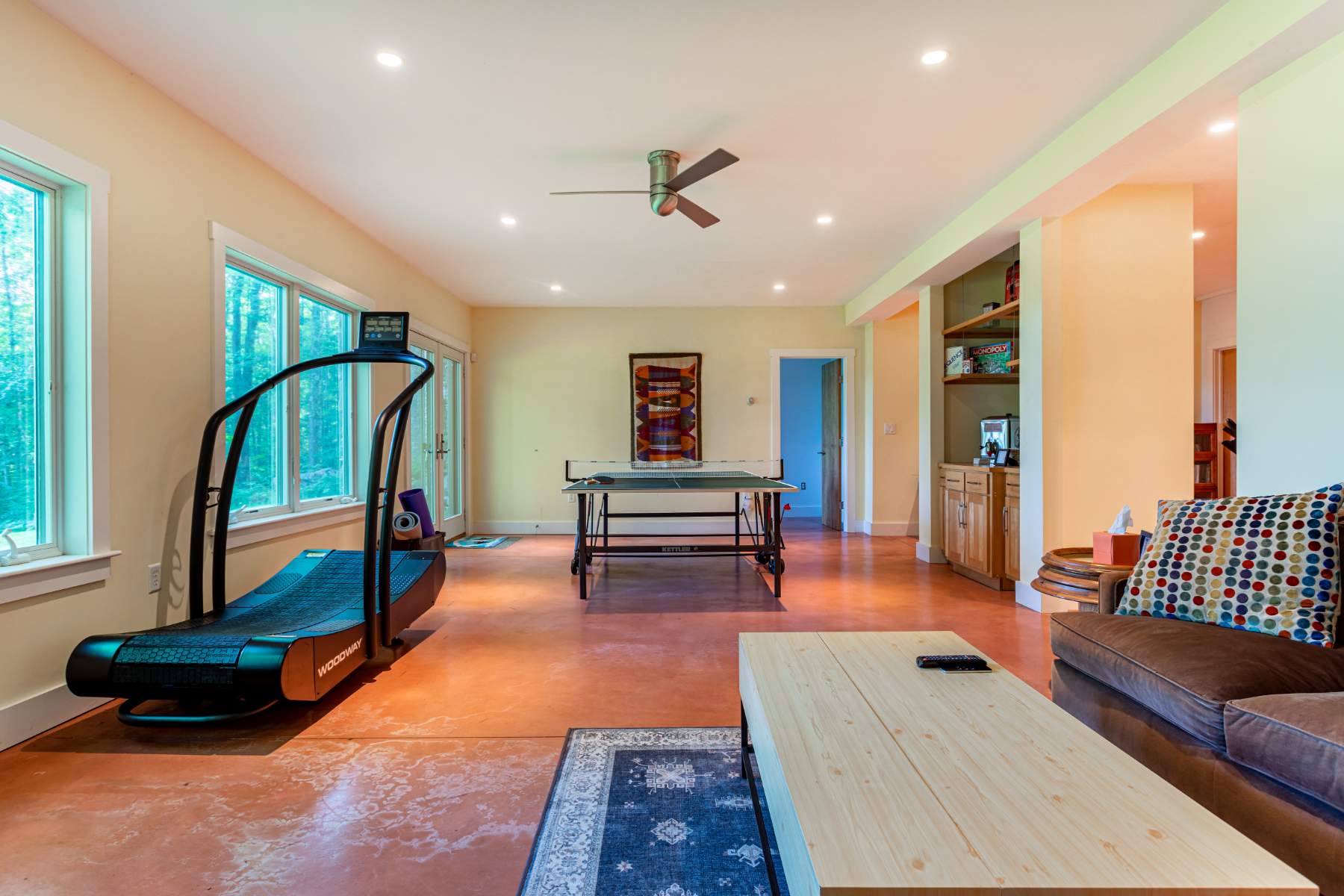 A spacious recreation room with a Kettler ping pong table, a black Woodway treadmill by large windows, and a cozy living area with a brown sofa and light wood coffee table, all on a polished orange concrete floor.