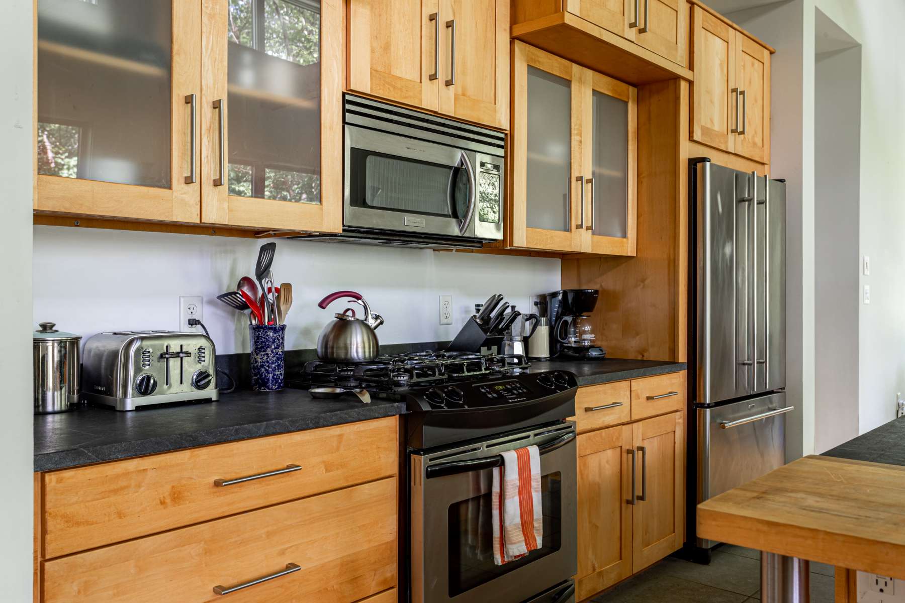 A detailed view of the kitchen counter shows a stainless steel gas range with an overhead microwave, surrounded by light wood cabinets with frosted glass inserts. Various kitchen essentials like a toaster, kettle, and coffee maker are neatly arranged on the black countertop.