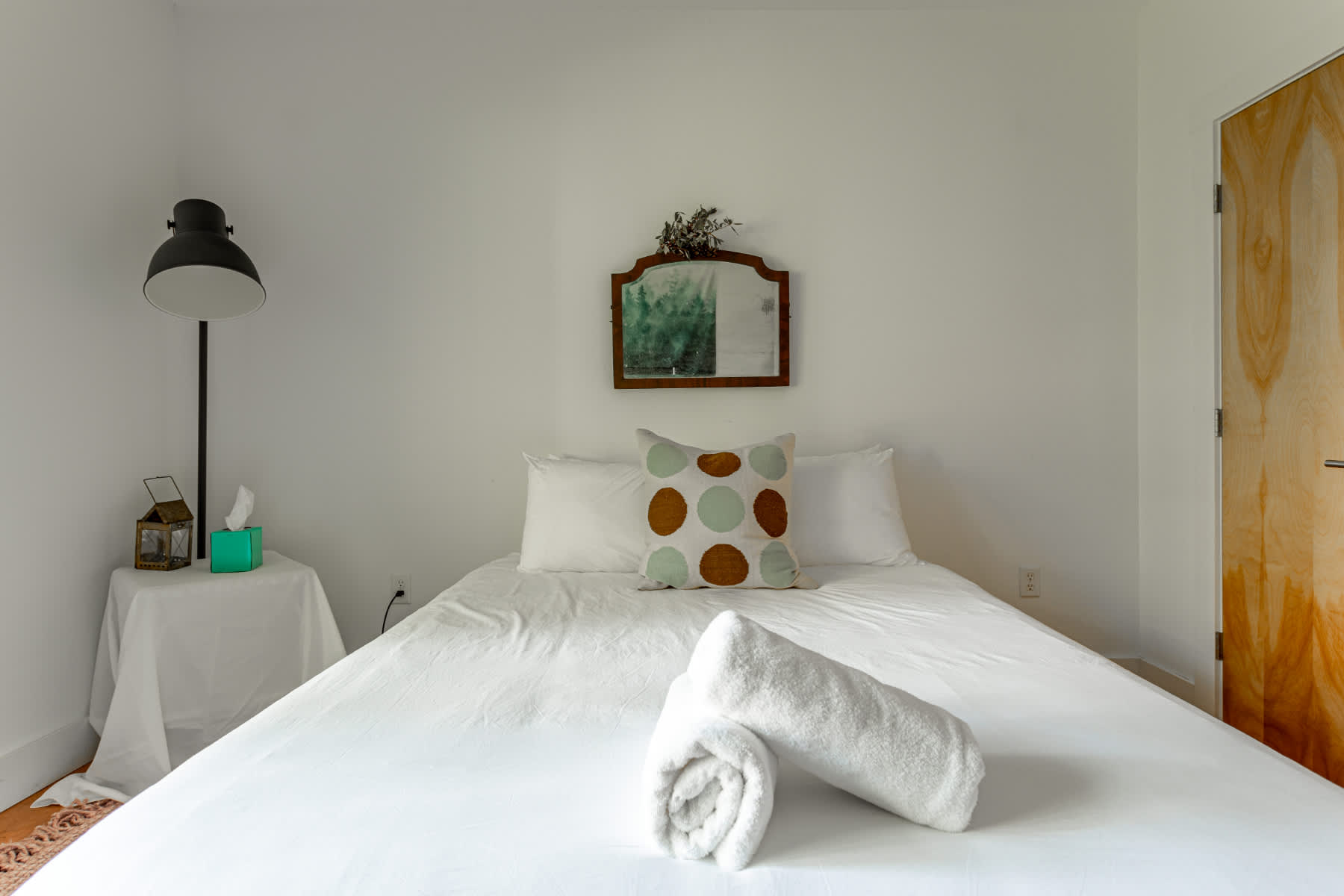 A bright bedroom features a queen-sized bed with crisp white linens and a polka-dot throw pillow, flanked by a black floor lamp and a small side table, with a rustic mirror hanging above the headboard.