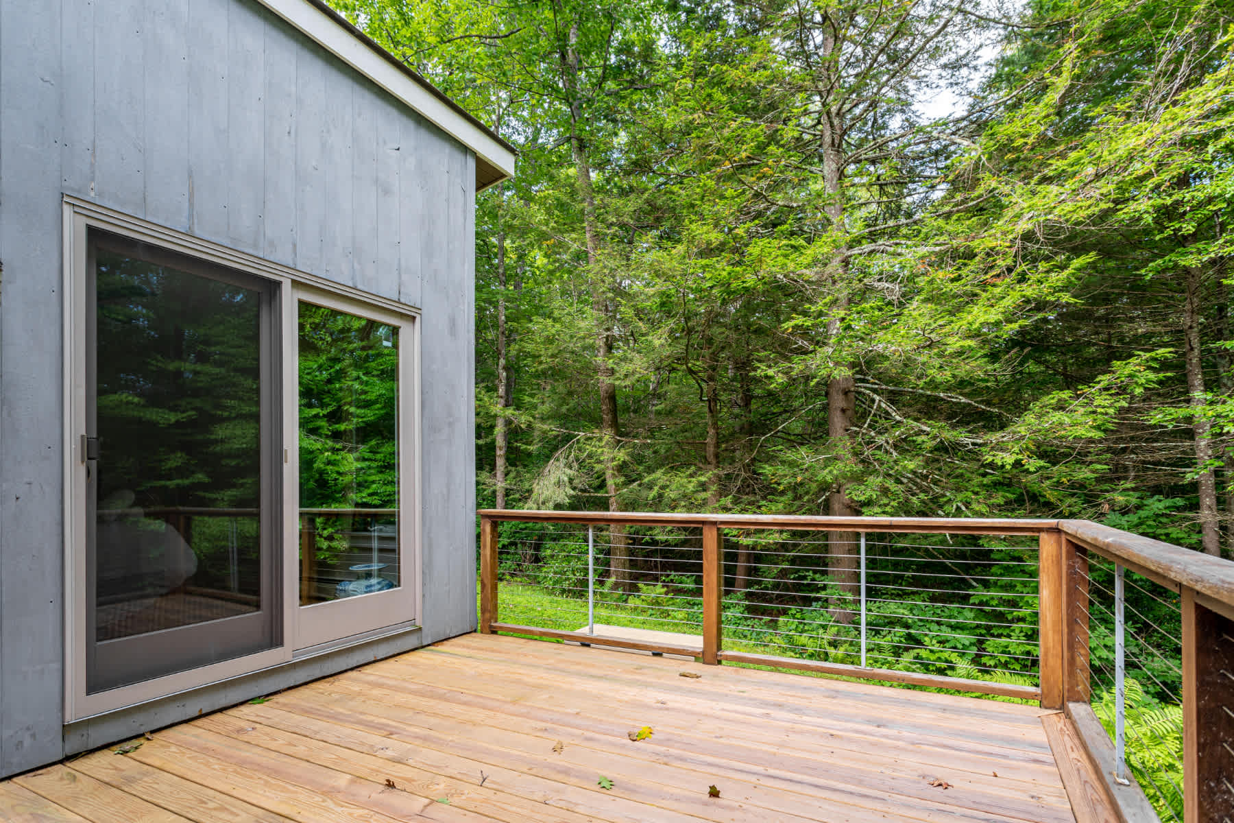 Spacious wooden deck surrounded by lush green trees, featuring a modern cable railing and a sliding glass door leading into the house.