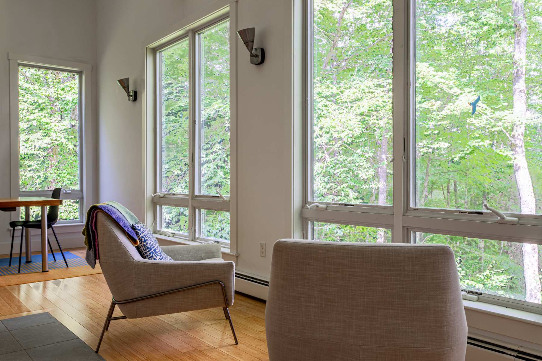 A bright and airy living area features light hardwood floors and tall, expansive windows that frame the lush green forest outside. Two modern light gray armchairs are strategically placed to offer comfortable seating with scenic views, adjacent to a dining area.