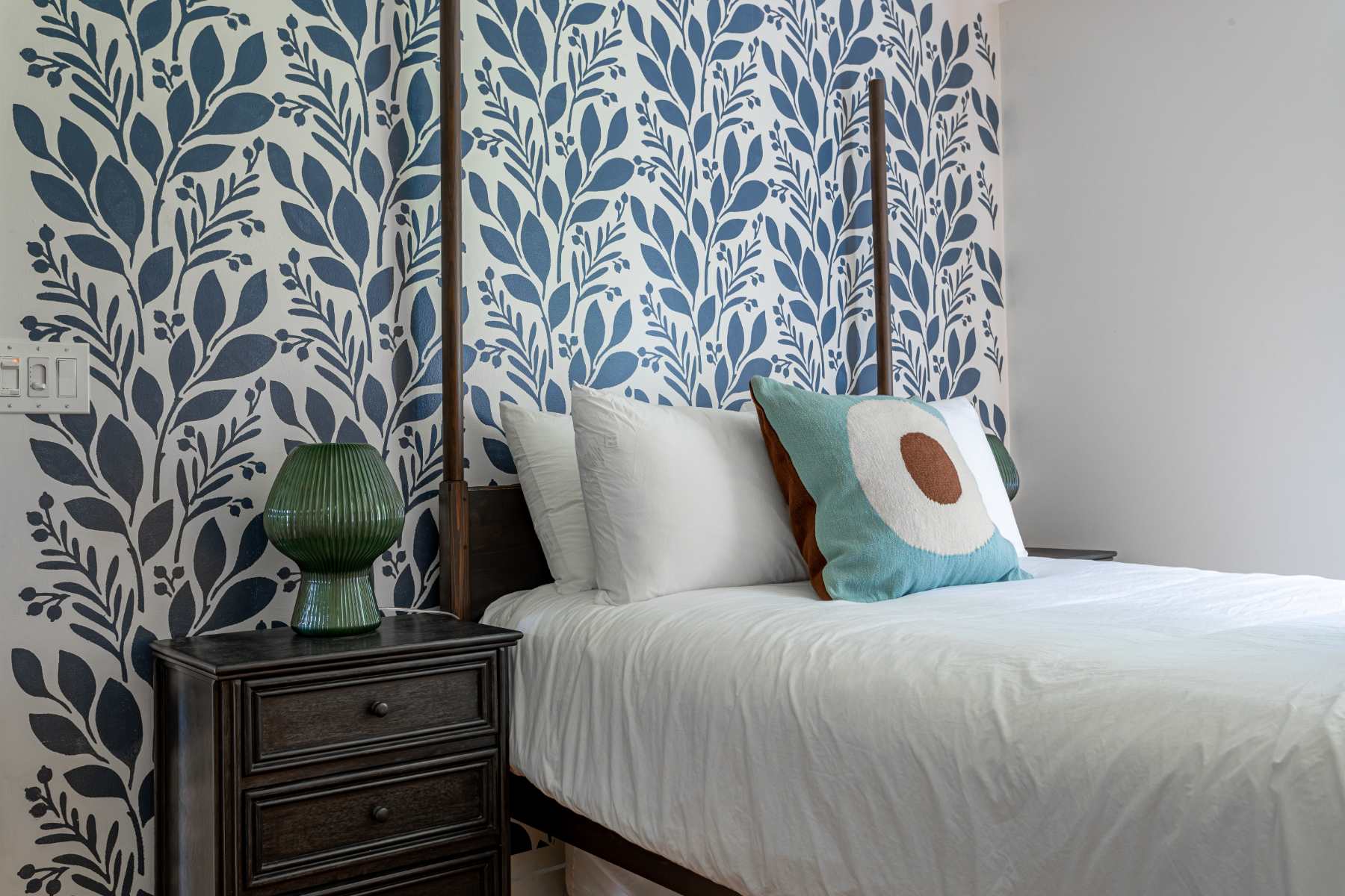 A close-up of a bedroom corner shows a dark wooden nightstand with a green ribbed lamp, next to a bed with white linens and a blue and brown patterned throw pillow. The wall behind features a bold blue and white botanical wallpaper.