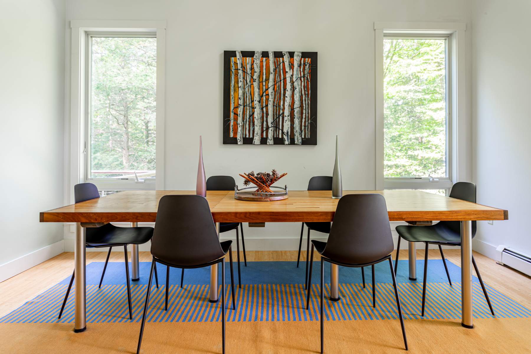 A bright dining room features a long wooden table with black chairs, set on a blue and orange striped rug. A modern birch tree painting hangs on the white wall between two large windows overlooking greenery.
