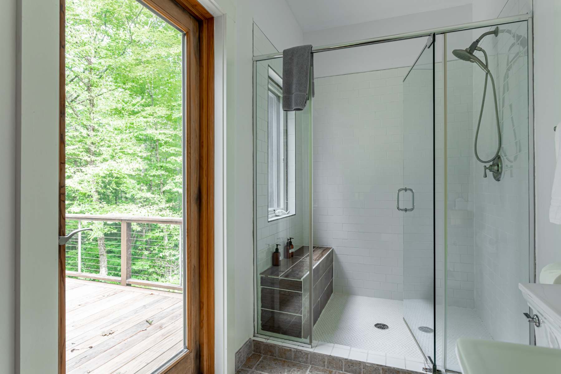 Modern walk-in shower with white subway tiles, a glass enclosure, and a built-in bench, featuring a window that provides natural light and a view of the outdoor deck and trees.
