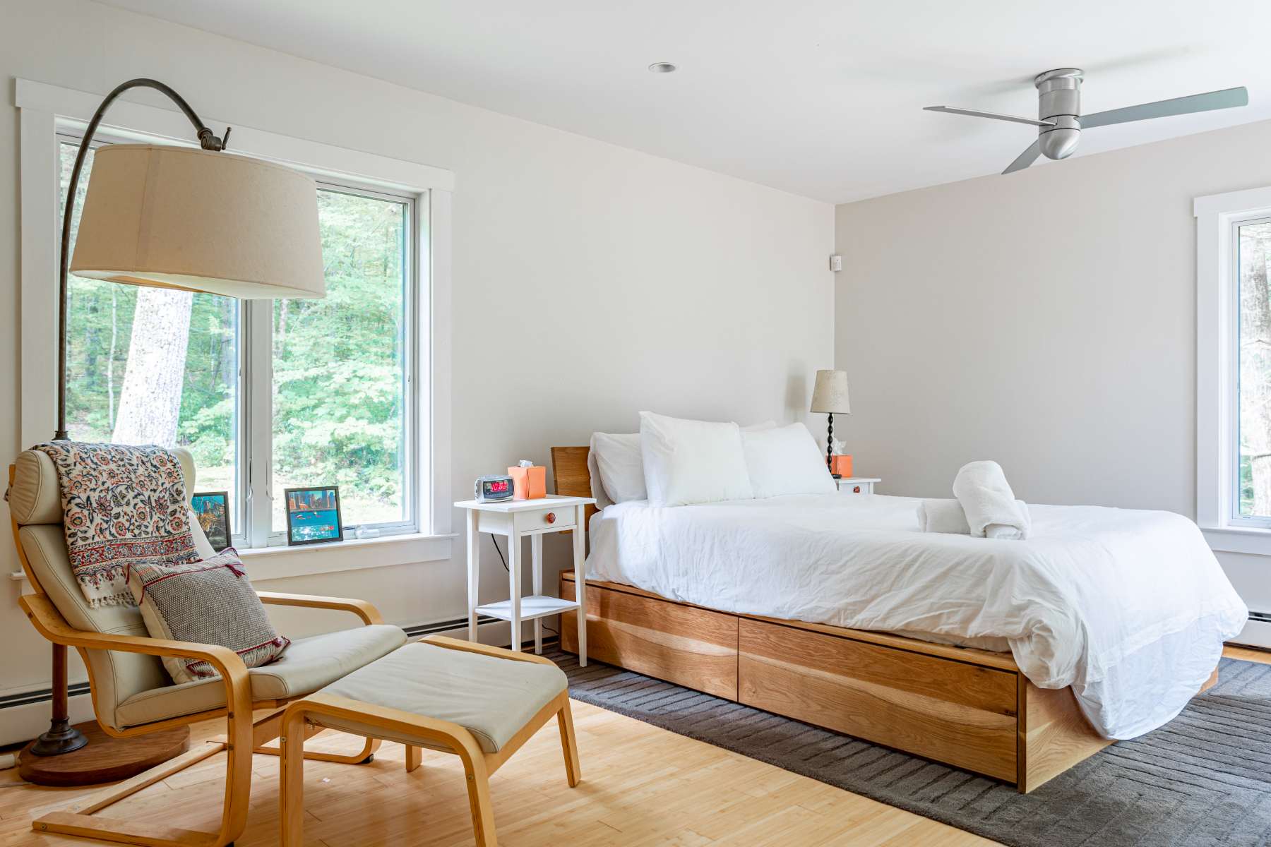 Bright bedroom with a white bed, light wood frame, and a white nightstand, featuring a large window overlooking lush greenery and a comfortable lounge chair with an ottoman.