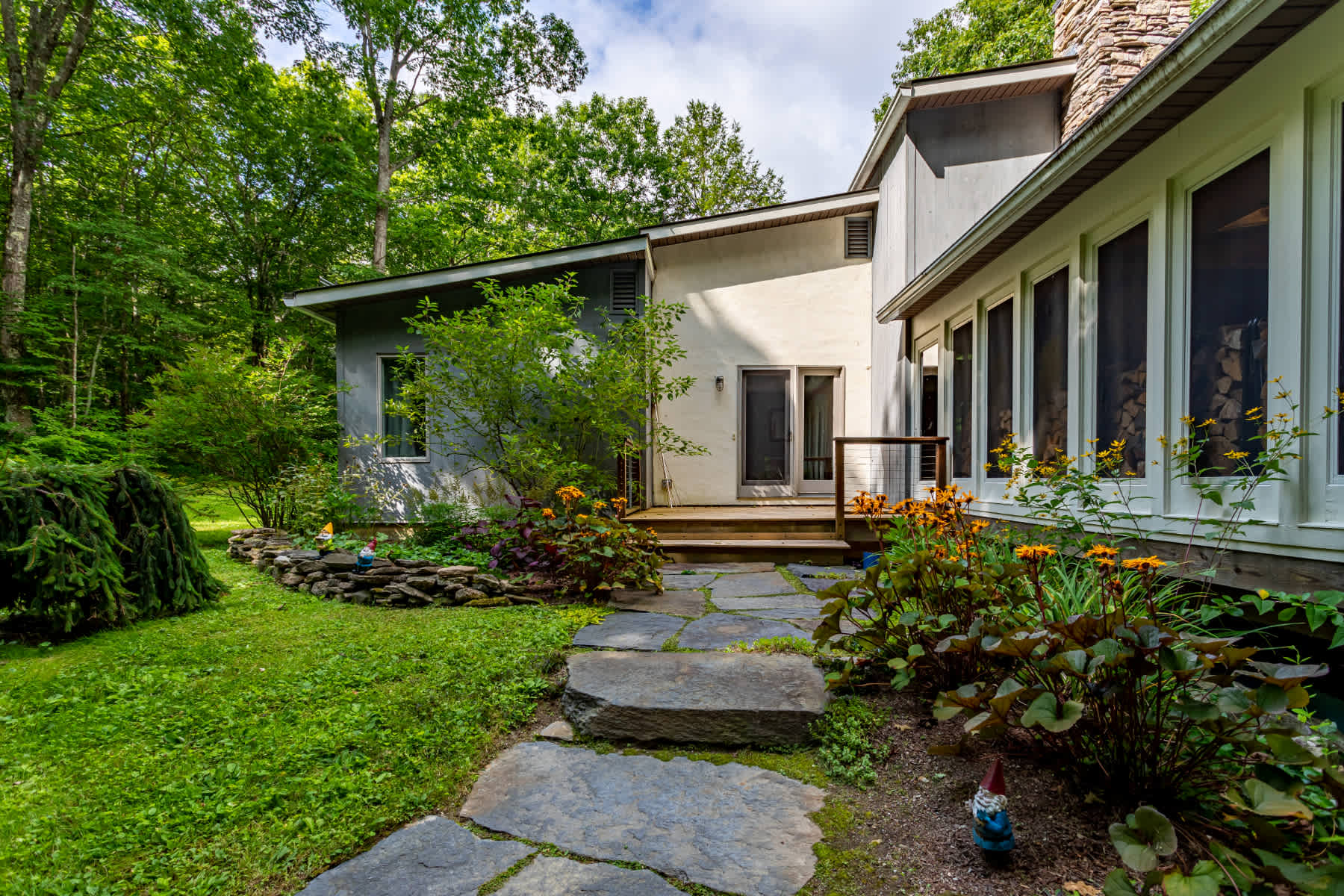 A stone pathway winds through a vibrant green lawn and garden beds with orange flowers, leading to a wooden deck and the back entrance of a multi-level house with light-colored siding.