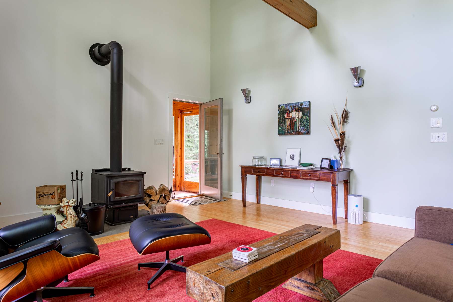 A cozy living room features a black wood-burning stove with a tall pipe, a black leather armchair, and a rustic wooden coffee table on a red area rug. A glass-paned door leads outside, while high white walls are adorned with artwork and exposed wooden beams.