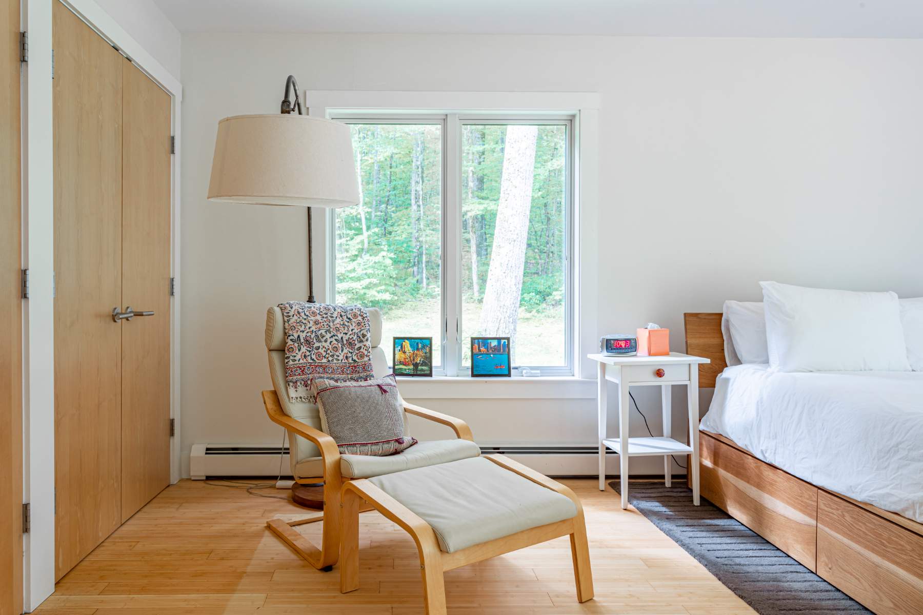 Cozy corner of a bedroom with a light wood lounge chair and ottoman, a large window offering views of trees, and a tall floor lamp, adjacent to a white nightstand and light wood closet doors.
