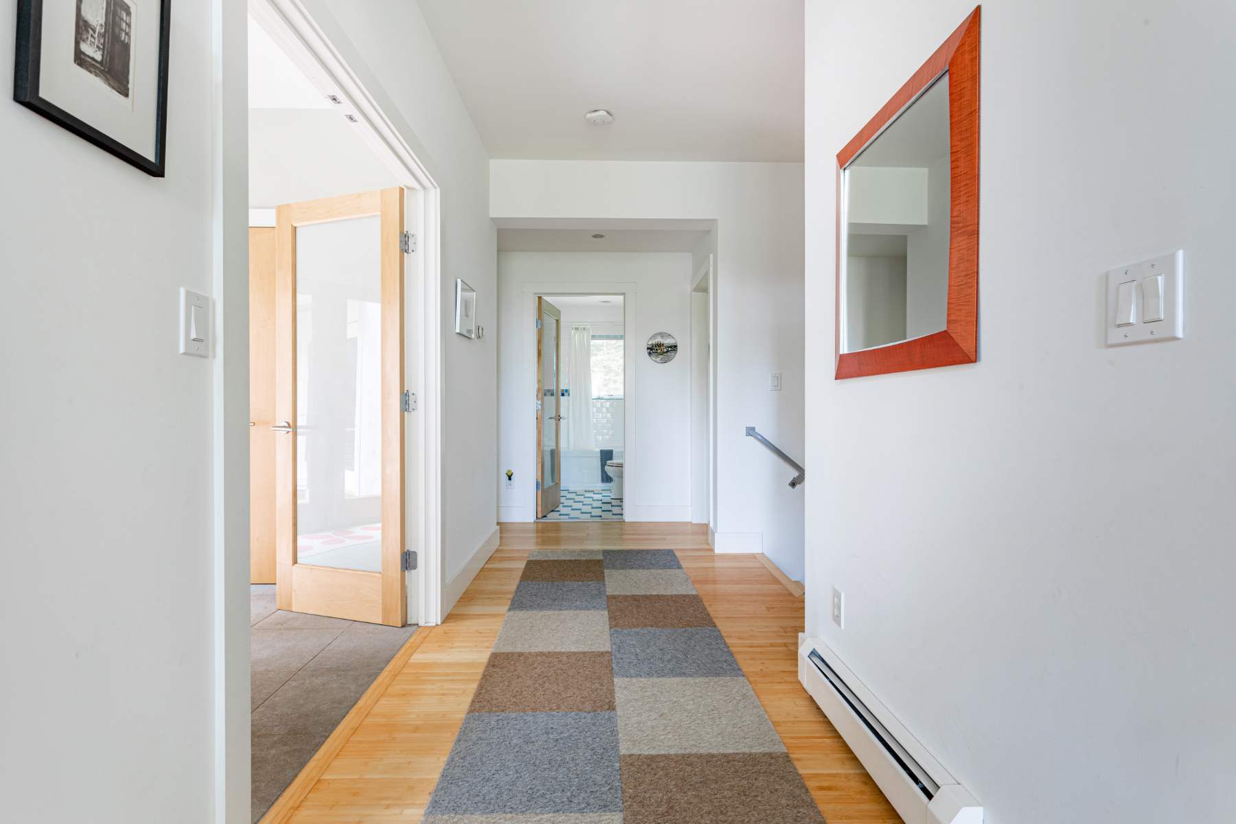 A bright hallway with light hardwood floors features a checkered runner rug leading towards another room. A mirror with a red frame hangs on the white wall, and a wooden door with frosted glass is visible on the left.