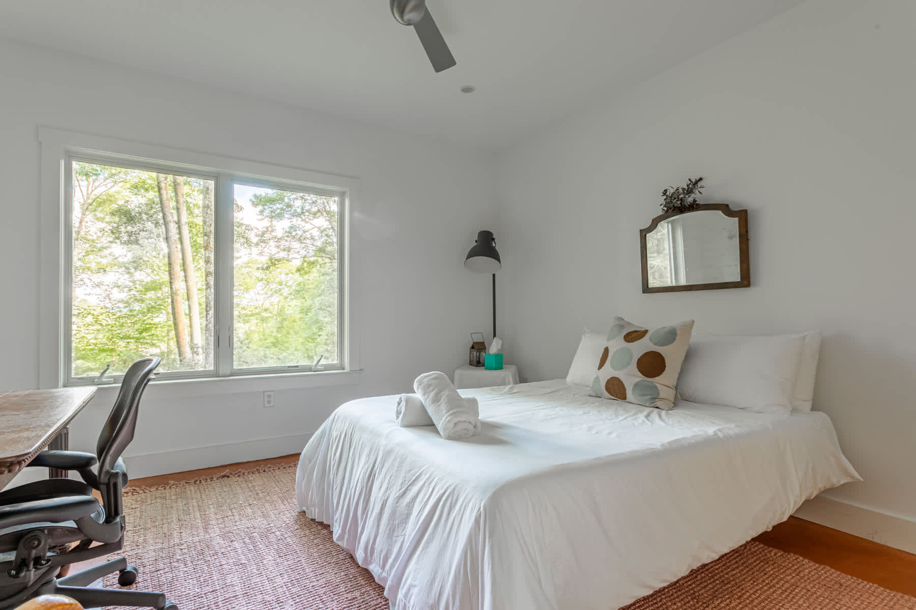 A serene bedroom with a queen-sized bed dressed in white, a wooden desk with an office chair by a large window overlooking lush greenery, and a ceiling fan for comfort.