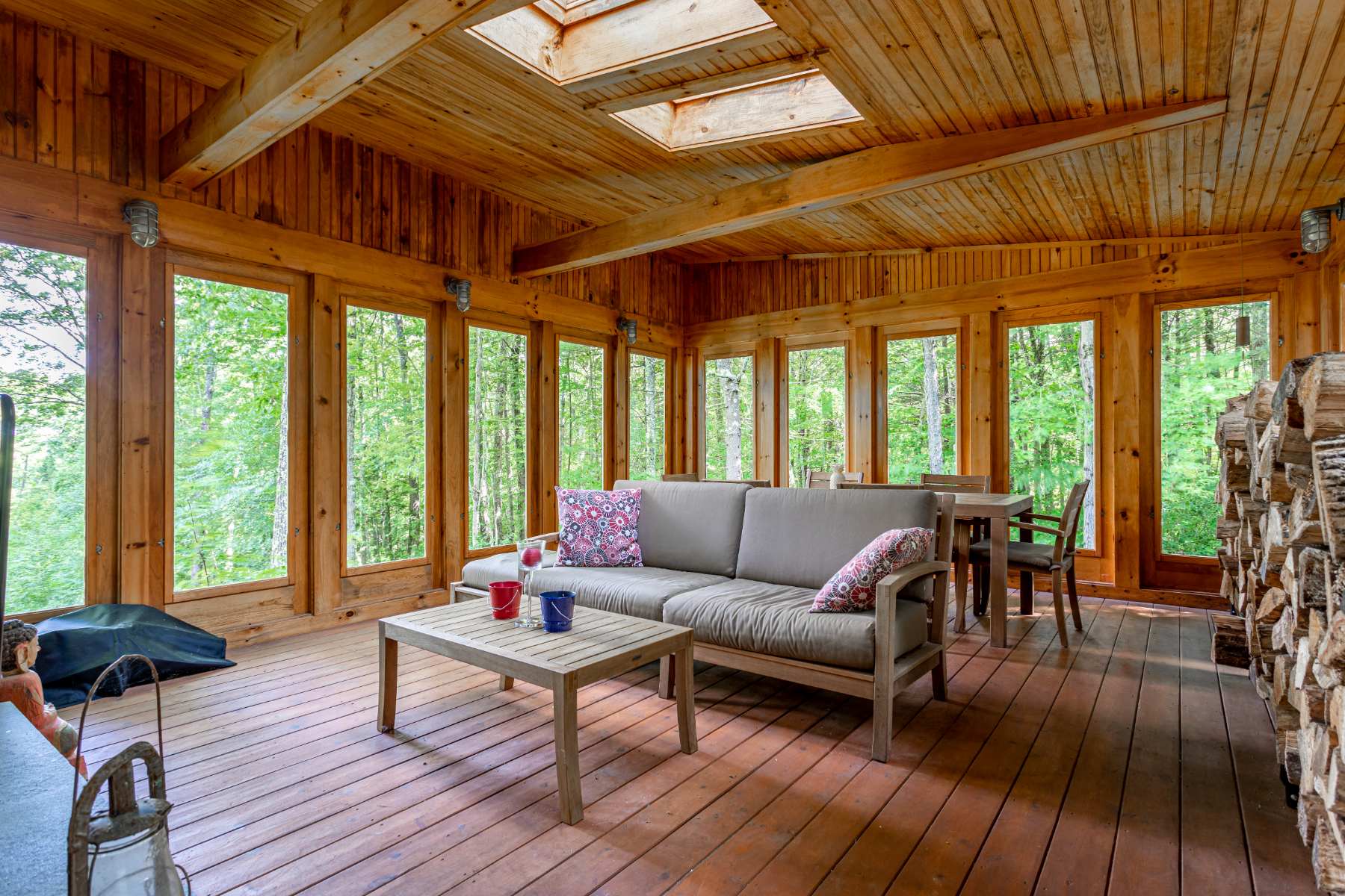 This rustic sunroom features natural wood paneling on the walls and ceiling, a skylight, and numerous large windows providing immersive views of the dense forest. It is furnished with a light gray outdoor sofa, a wooden coffee table, and a dining table, with stacked firewood visible in the corner.