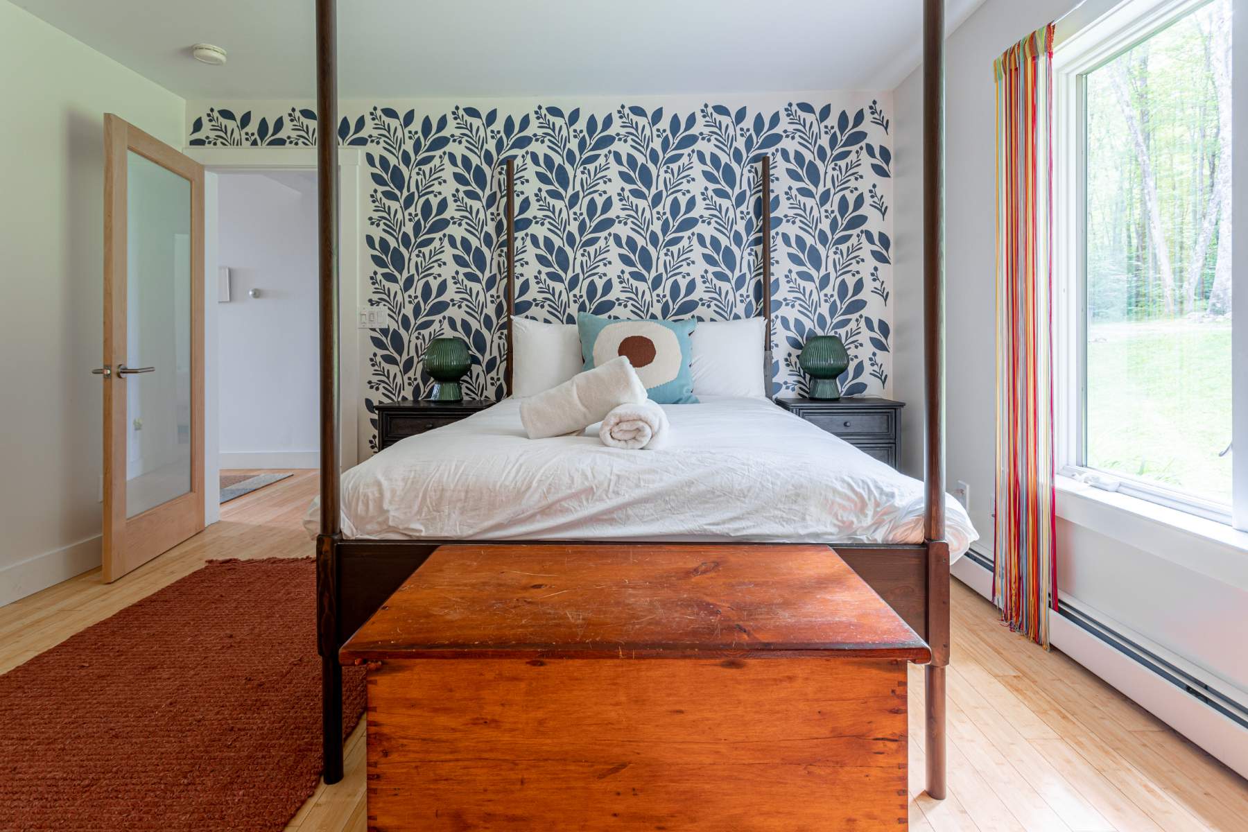 A spacious bedroom features a dark wooden four-poster bed with white linens and a blue botanical wallpaper accent wall. A rustic wooden trunk sits at the foot of the bed, and a large window with colorful striped curtains offers a view of the outdoors.