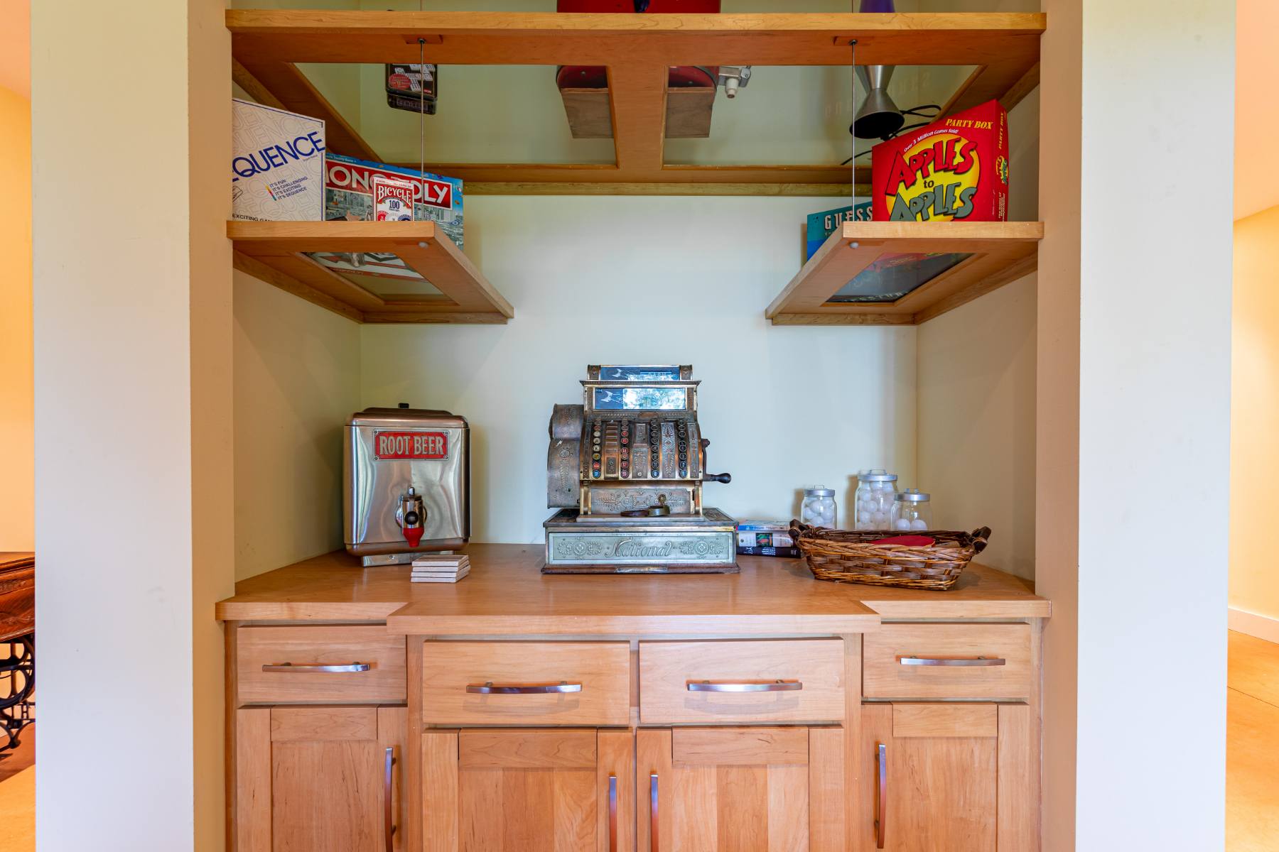 A built-in wooden cabinet features open shelves displaying board games like Sequence and Apples to Apples, with an antique cash register and a vintage root beer dispenser on the countertop below.