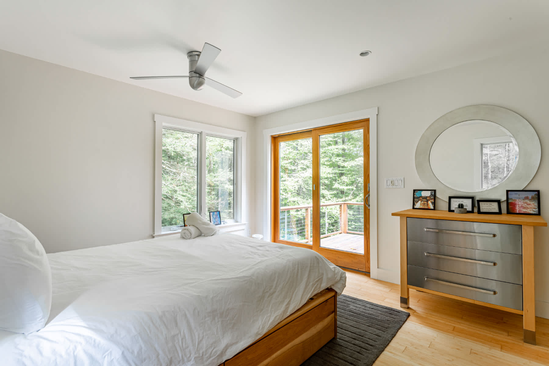 Modern bedroom featuring a white bed, a dresser with metal drawers and a round mirror, and a sliding glass door leading to an outdoor deck with views of surrounding trees.