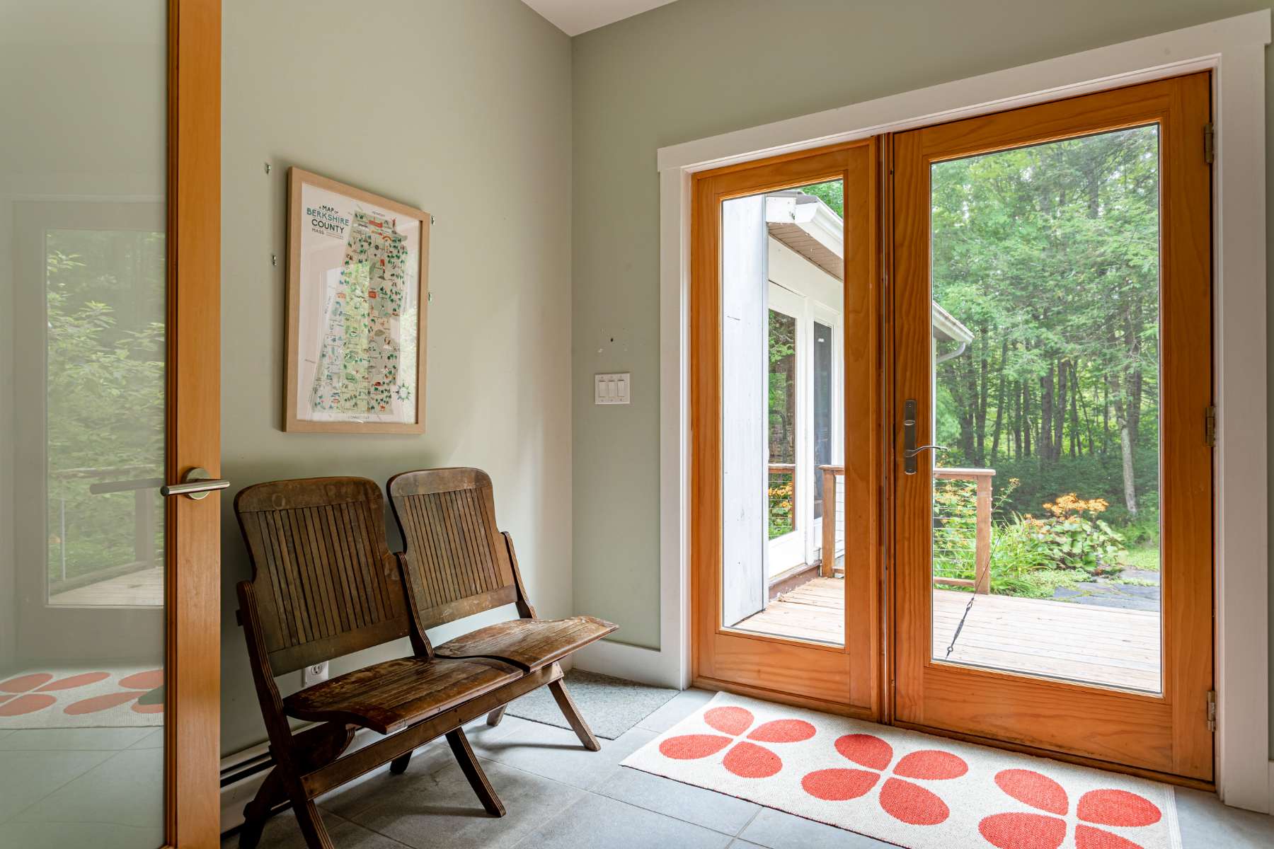 A welcoming entryway features light green walls and a grey tiled floor, with two rustic wooden folding chairs against one wall. Double glass doors open to an outdoor deck, and a red and white floral rug adds a cheerful accent.