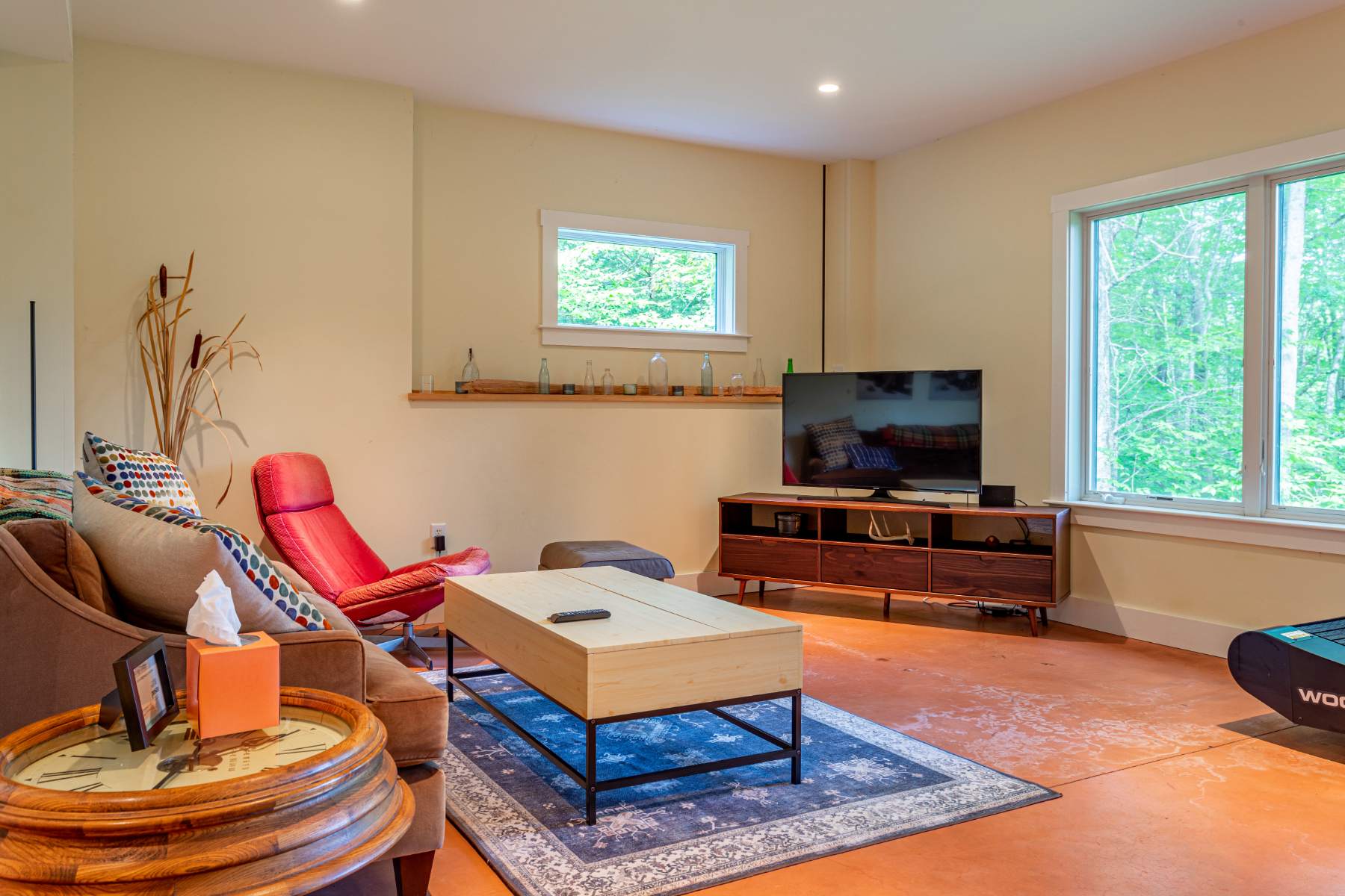 A cozy living area with light yellow walls and large windows provides abundant natural light and views of the surrounding trees. It is furnished with a comfortable brown sofa, a vibrant red lounge chair, a light wood coffee table, and a TV on a wooden console, all resting on a blue patterned rug over a reddish-brown floor.