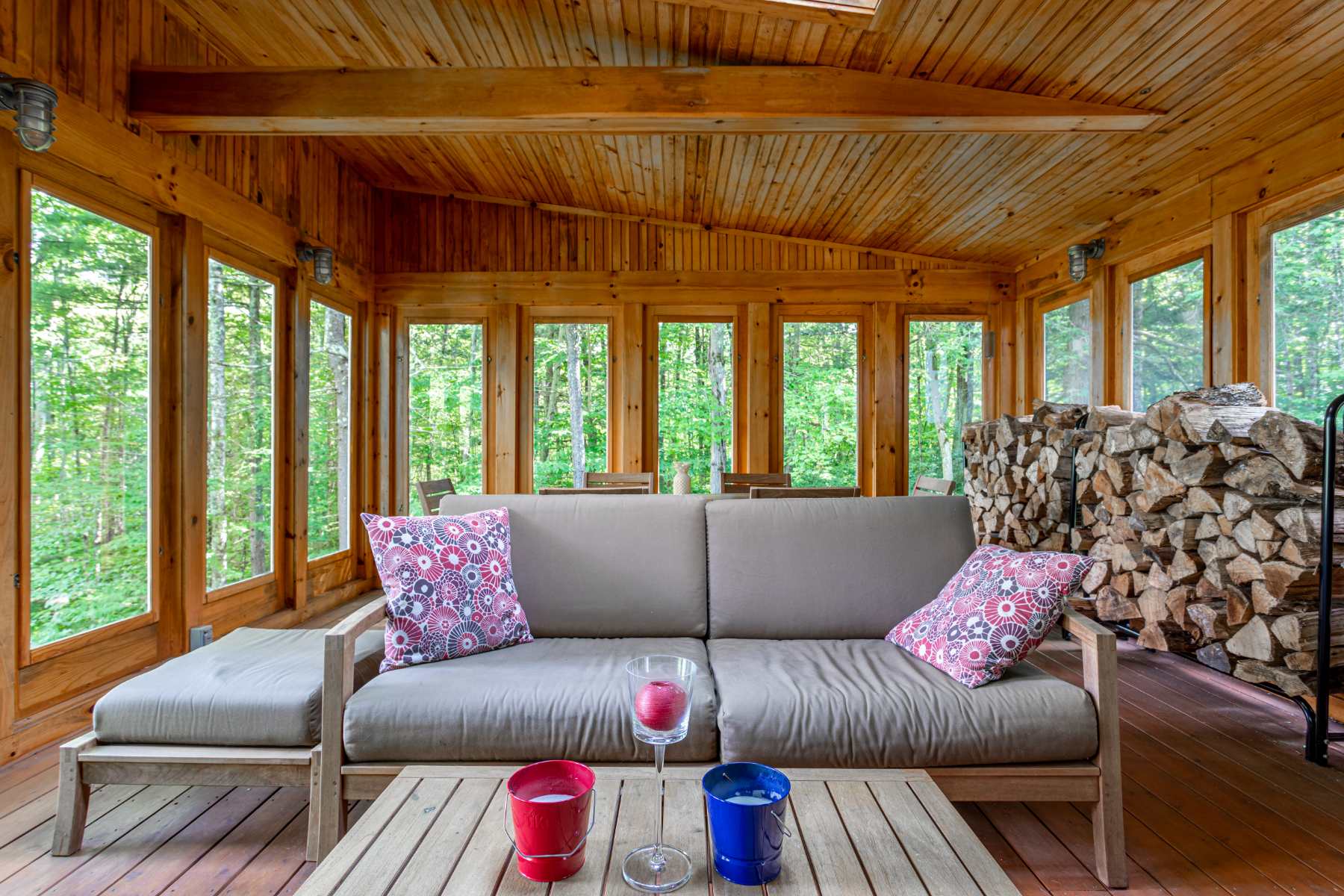 A cozy sunroom features a light brown sofa with colorful throw pillows, a wooden coffee table with candles, and large windows offering a lush green forest view. The room has natural wood paneling on the walls and ceiling, creating a rustic, inviting atmosphere, with stacked firewood in the corner.