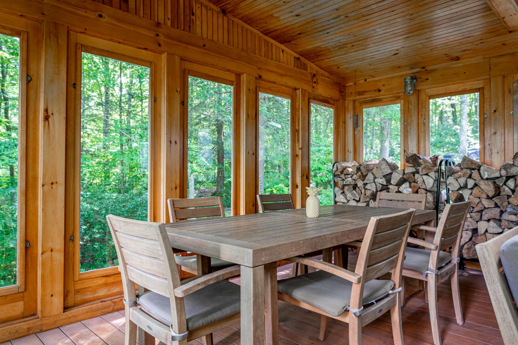 A bright dining area in a sunroom showcases a wooden dining table with six chairs, adorned with a small pineapple decoration. Large windows line the wooden-paneled walls, providing expansive views of the surrounding green forest and stacked firewood in the corner.