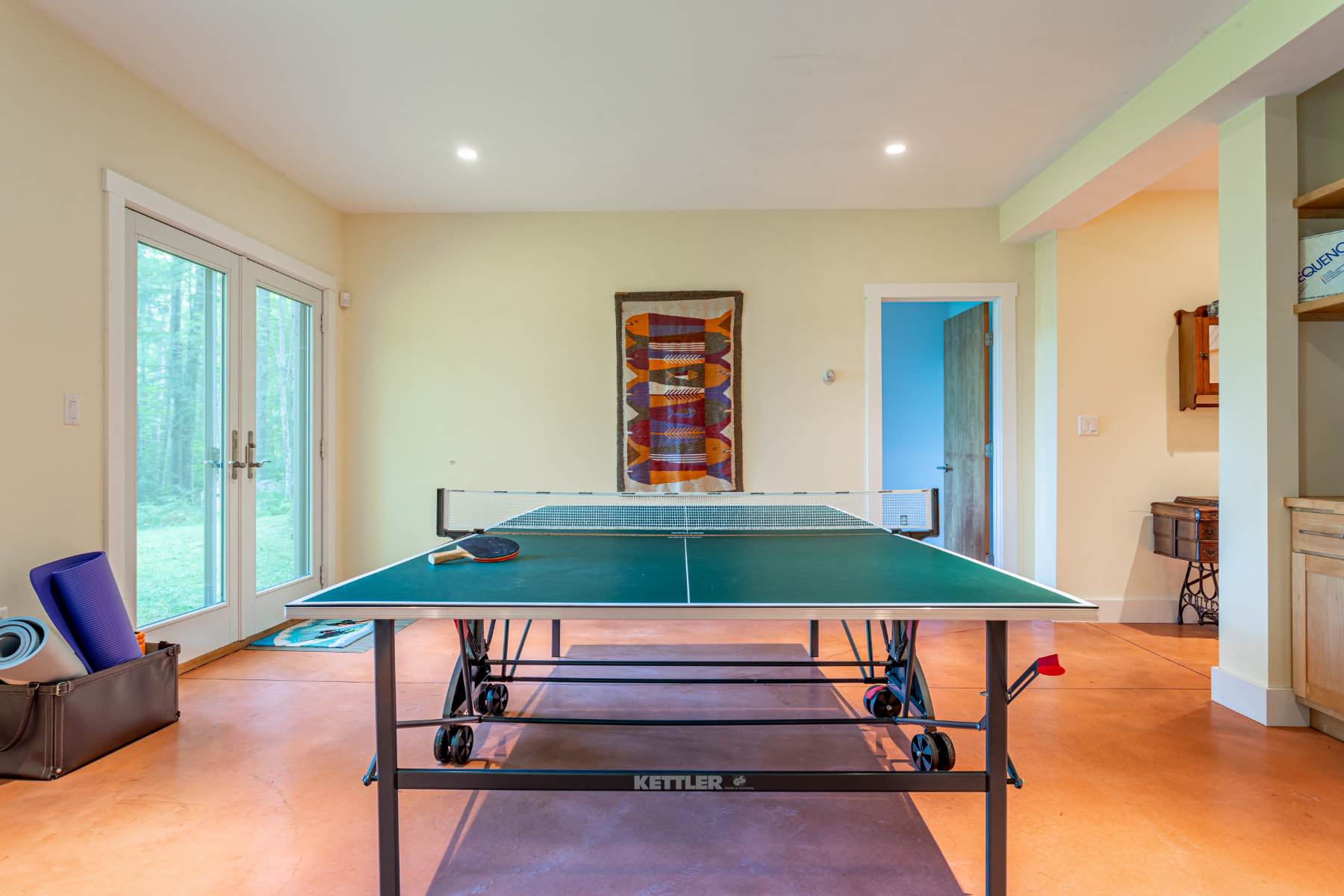 A bright game room features a green Kettler ping pong table with paddles and balls, set on a polished orange concrete floor, with double glass doors leading outside and a colorful woven tapestry on the wall.