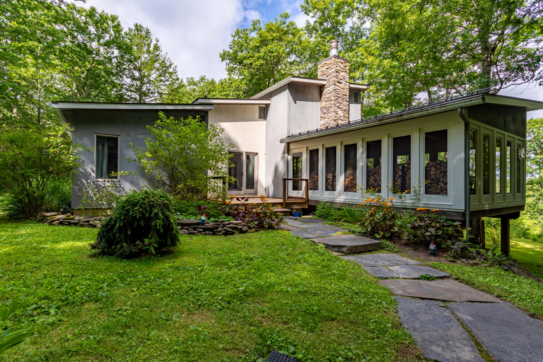 The back of a modern house with light-colored siding and a prominent stone chimney, surrounded by a lush green lawn and mature trees, with a stone path leading to a wooden deck.