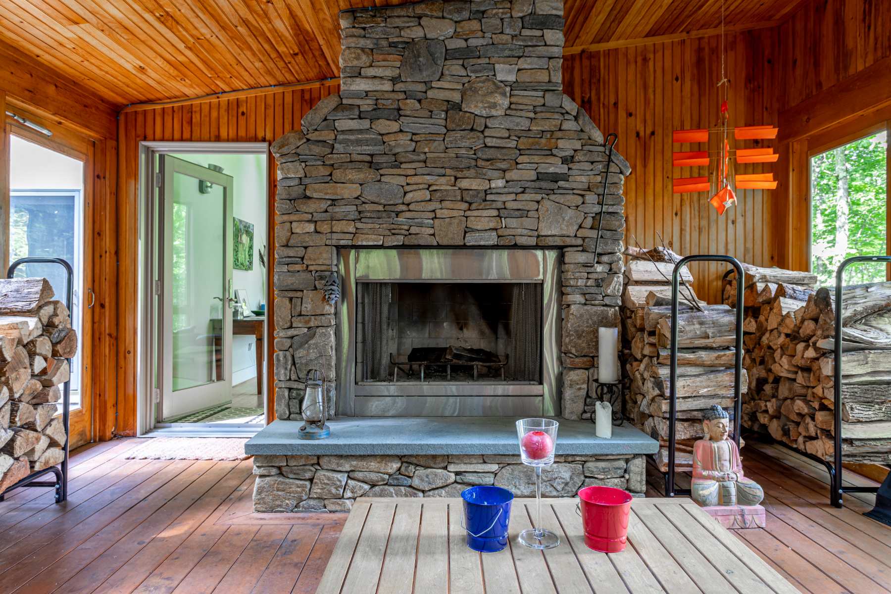 A cozy sunroom with natural wood walls and ceiling is centered around a large, impressive stone fireplace with a metal insert. Stacked firewood is neatly arranged on both sides of the hearth, and a wooden coffee table with colorful cups sits in the foreground.