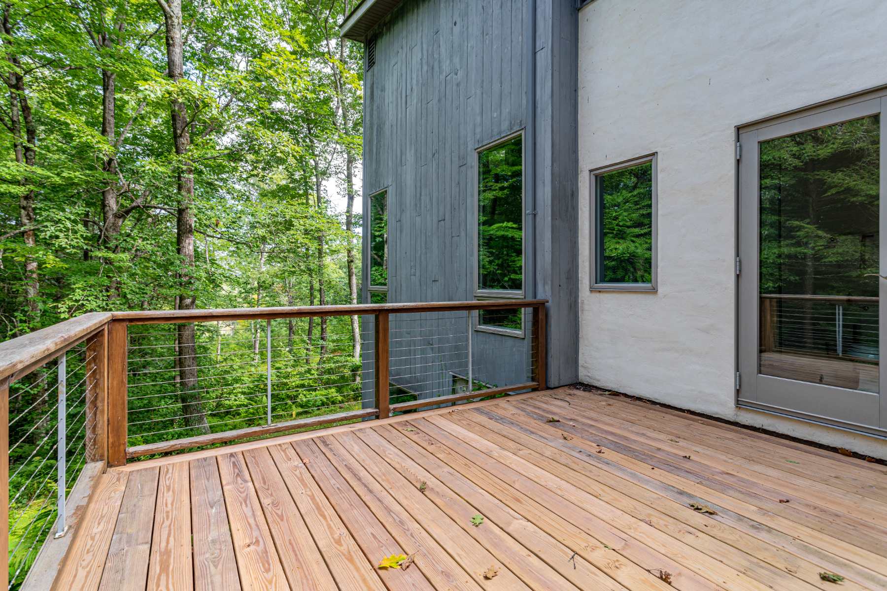Expansive wooden deck with a cable railing, offering views of dense green trees and showcasing the modern exterior of the house with gray siding and white stucco sections.
