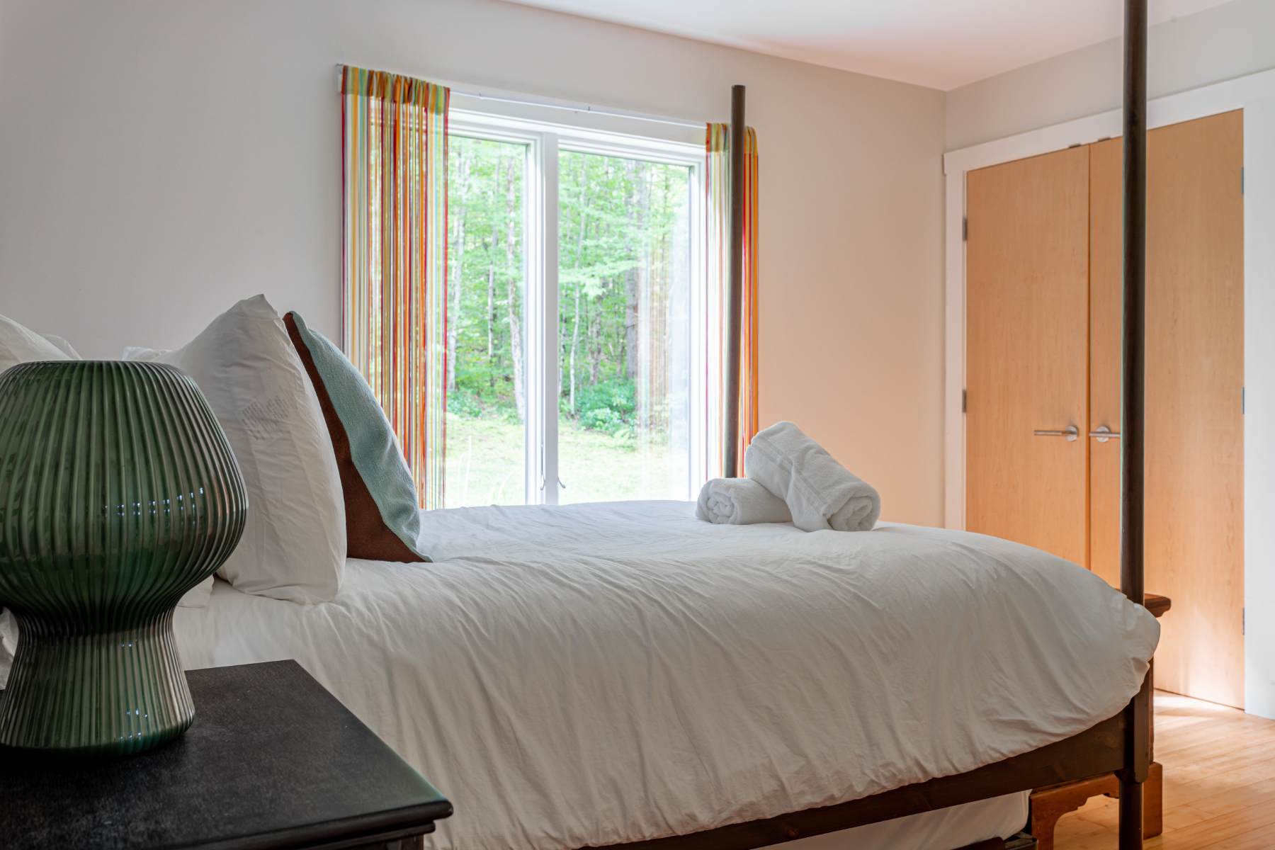 A bright bedroom corner shows a bed with white linens and a blue throw pillow, next to a dark nightstand with a green ribbed lamp. A large window with colorful striped curtains provides a view of lush greenery outside.