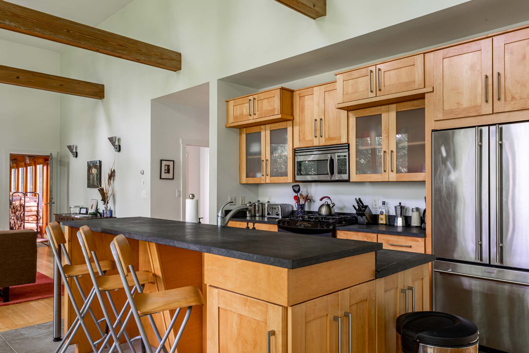 A modern kitchen features light wood cabinetry, black countertops, and a central island with bar seating and three wooden stools. Stainless steel appliances, including a refrigerator and microwave, are integrated into the design, with a view into the living area.
