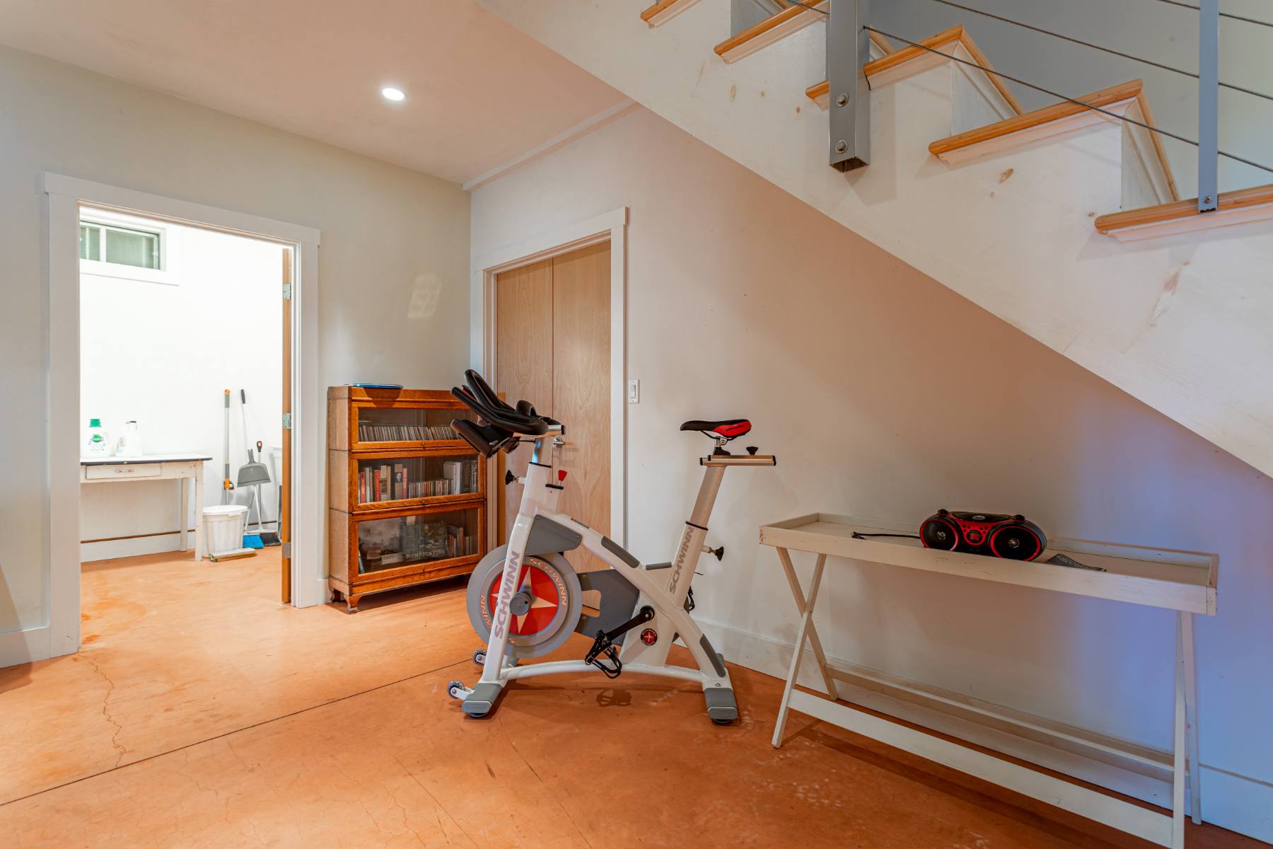 Recreation area featuring a white spin bike, a small wooden bookshelf, and a set of stairs with modern railings, with a laundry room visible through an open doorway.