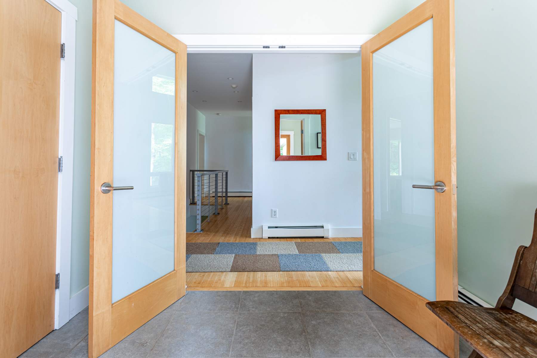 Looking from an entryway with grey tiled floors through double frosted glass doors into a hallway with hardwood floors and a checkered runner rug. A mirror with a red frame is visible on the far wall.