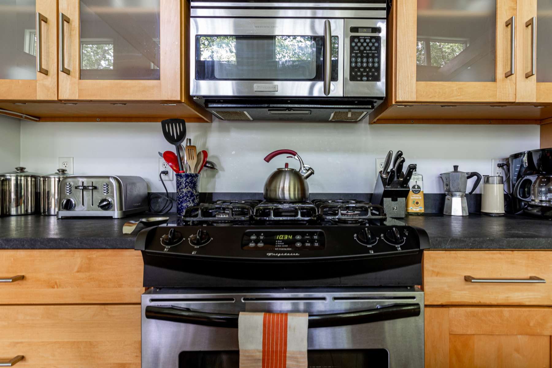A close-up of the kitchen's cooking area features a stainless steel gas range with a digital clock, topped by a metal kettle and surrounded by various cooking utensils. The light wood cabinetry and black countertop are visible, with a microwave above the stove.