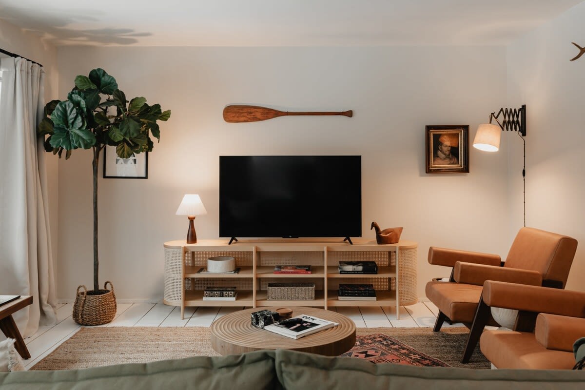 A cozy living room featuring a large TV on a light wood and cane media console, flanked by a tall fiddle leaf fig plant and a brown leather armchair, with a decorative oar mounted above the TV.