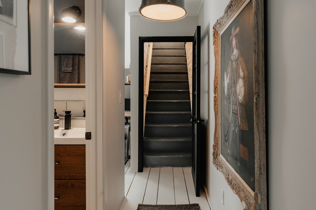 Bright hallway with white walls and painted white floorboards, leading to a bathroom on the left and a dark staircase on the right. A large framed painting hangs on the wall, adding character.