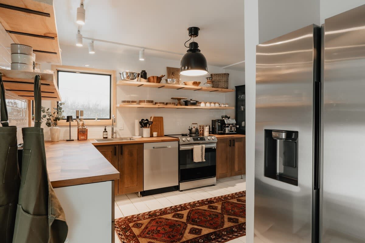 A spacious kitchen with stainless steel appliances including a refrigerator and range, wood cabinetry, herringbone countertops, and open shelving, featuring a patterned area rug on white tiled floors.