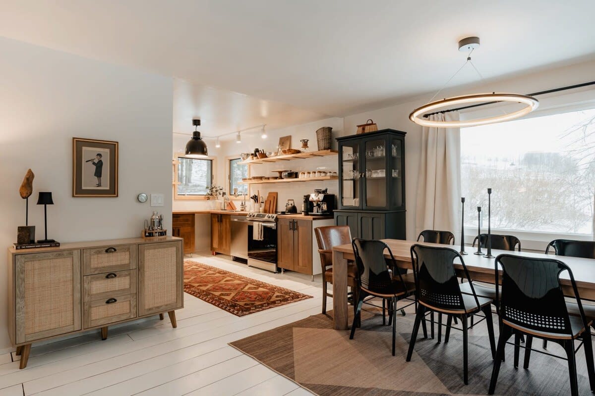 An open-concept space showing a dining area with a light wood table and black chairs, leading into a kitchen with wood countertops, white subway tile backsplash, and open shelving, all under a modern circular chandelier.