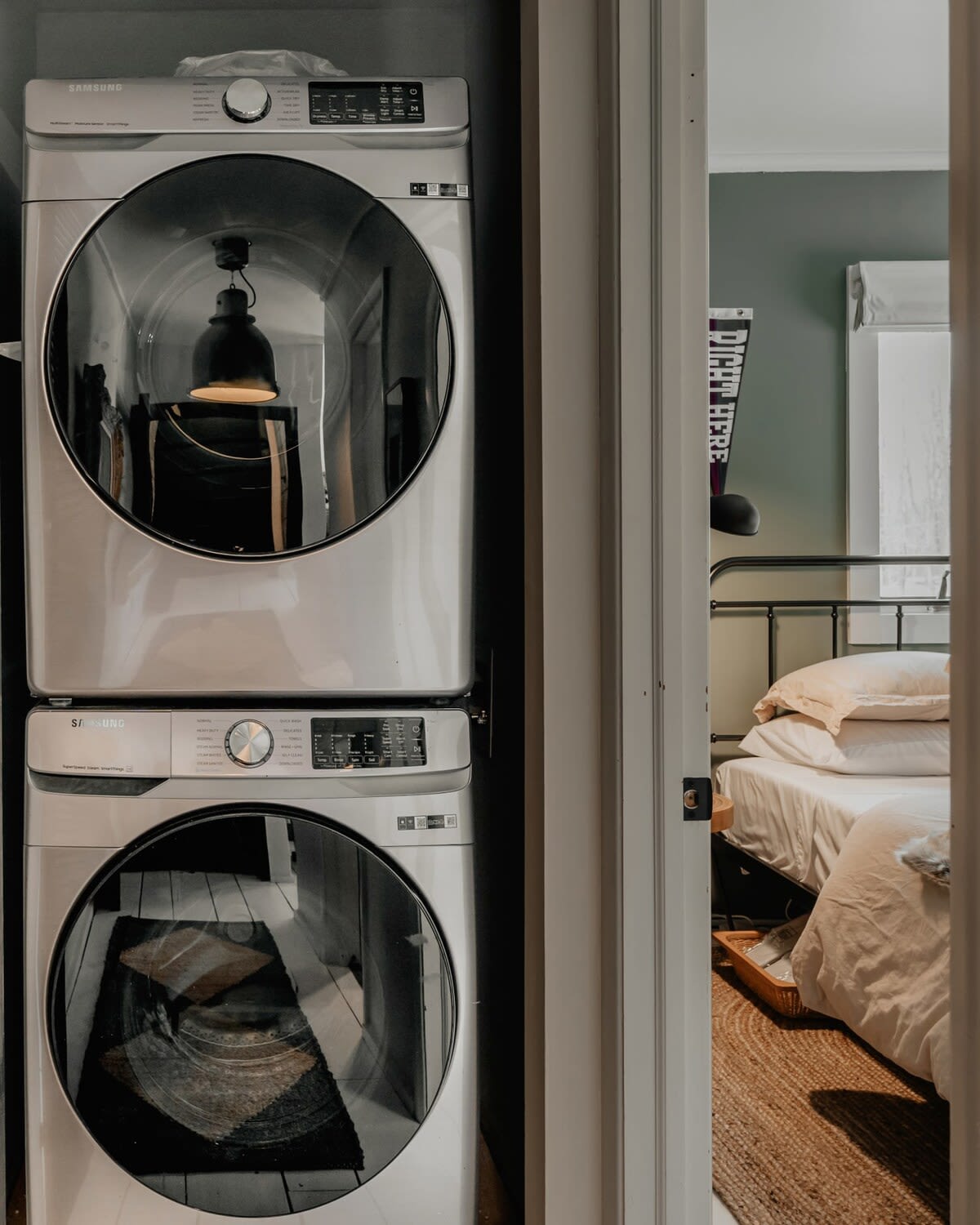 Compact laundry area featuring a stacked white Samsung washer and dryer unit. A glimpse of a bedroom with dark green walls is visible through the open doorway, suggesting proximity to living spaces.