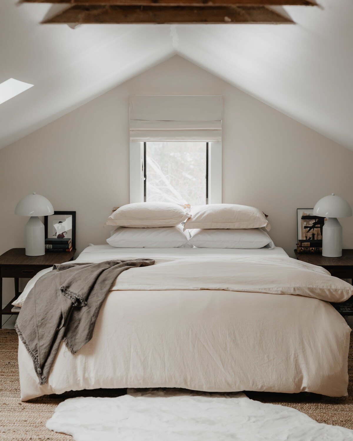 A cozy attic bedroom features a sloped white ceiling and a bed with light beige bedding, flanked by dark wood nightstands with white mushroom-shaped lamps. A small window with a white roman blind is centered above the bed, with exposed wood beams overhead.