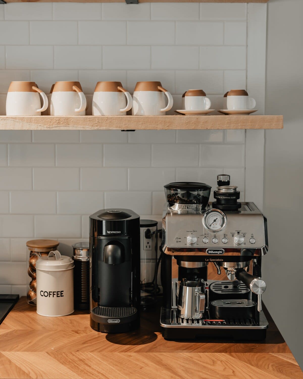 A close-up of a coffee station in the kitchen, featuring a Nespresso machine and a De'Longhi espresso machine on a herringbone wood countertop, with various coffee cups on an open shelf above.