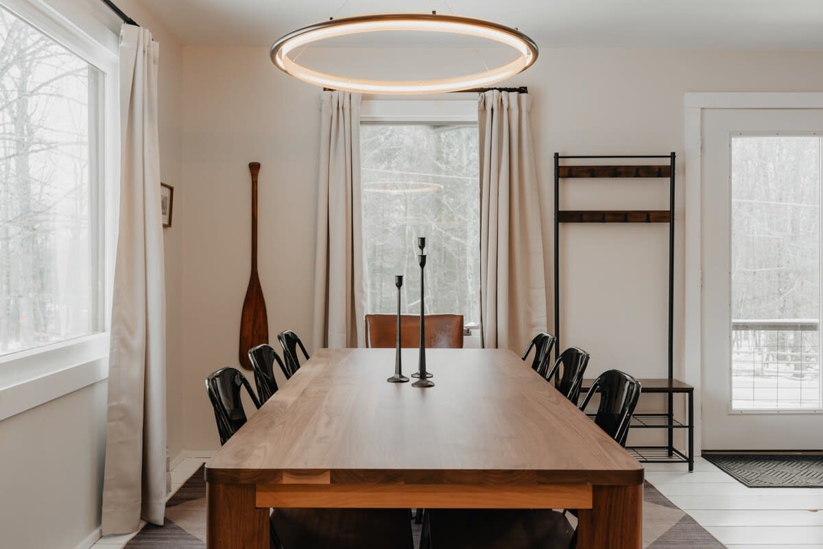 A spacious dining room features a long wooden dining table with black chairs, illuminated by a modern circular chandelier. Large windows with light curtains offer views of the surrounding nature.