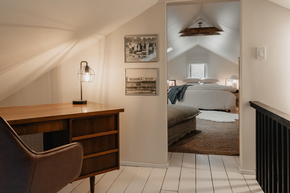 Bright attic landing area with white walls and painted white floorboards, featuring a wooden desk and chair. It leads into a spacious bedroom with an angled ceiling, offering a dedicated workspace.