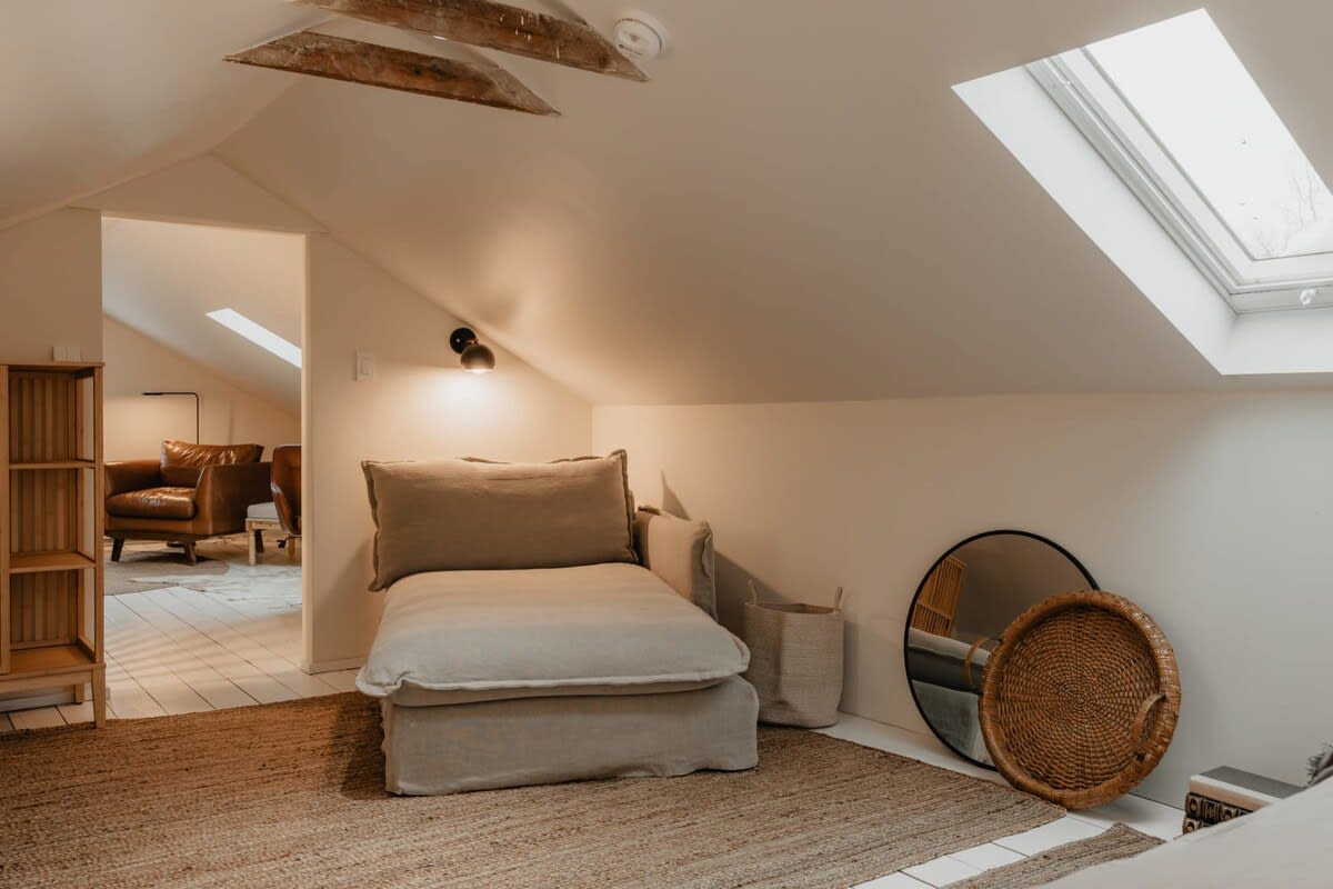 Bright attic lounge area with white walls and angled ceilings, featuring a comfortable beige chaise lounge and a round black-framed mirror. A jute rug covers the white floorboards, creating a cozy retreat.