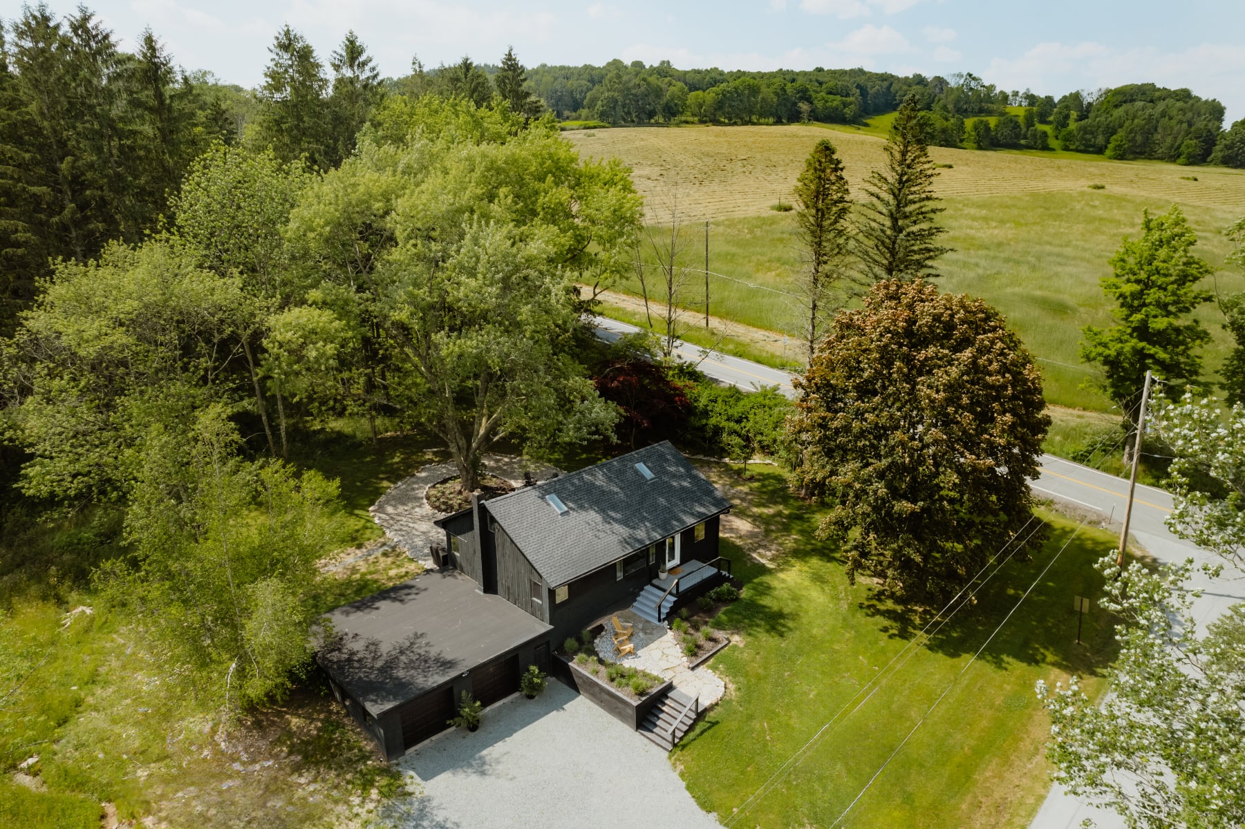 An aerial view showcases the black-sided cabin nestled among lush green trees and a sprawling lawn, with a gravel driveway and a road visible in the distance, highlighting its rural setting.