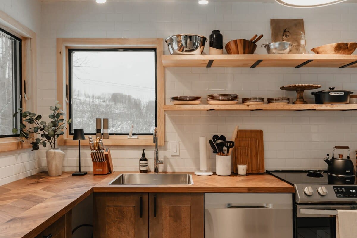 A well-lit kitchen corner featuring a stainless steel sink set into herringbone wood countertops, white subway tile backsplash, open wood shelving with dishes, and a window overlooking a snowy landscape.