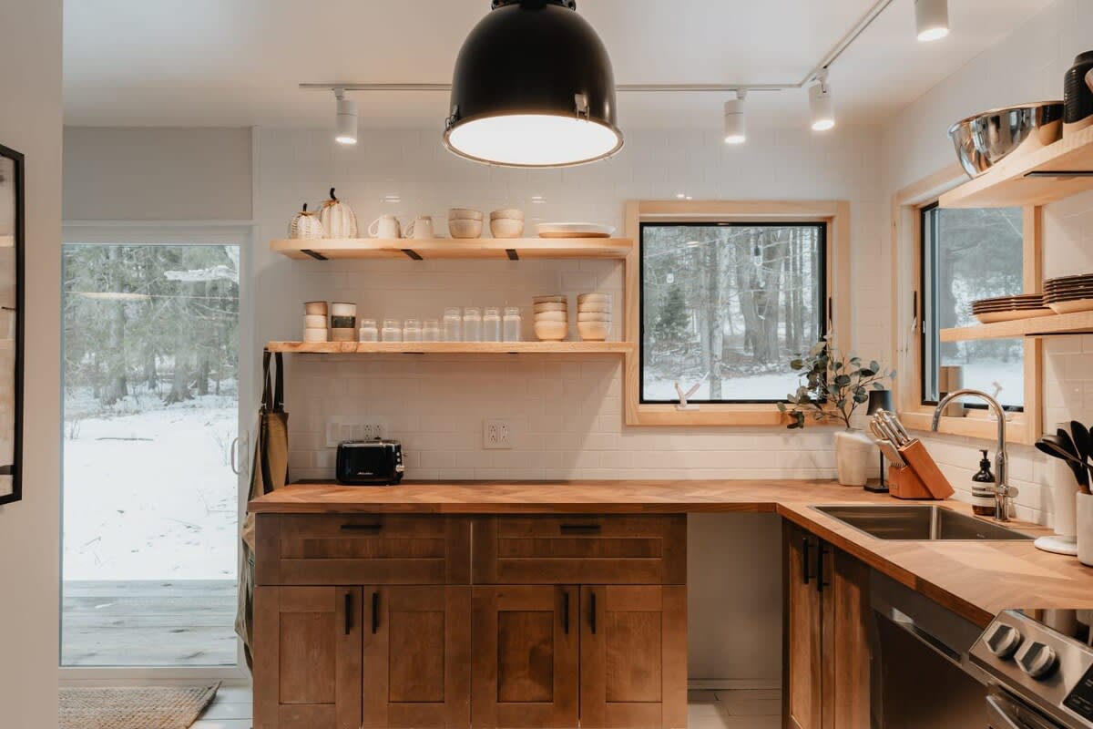 A view of the kitchen with warm wood cabinetry, herringbone wood countertops, white subway tile backsplash, and open shelving, illuminated by track lighting and a large black pendant light, with a window showing a snowy view.