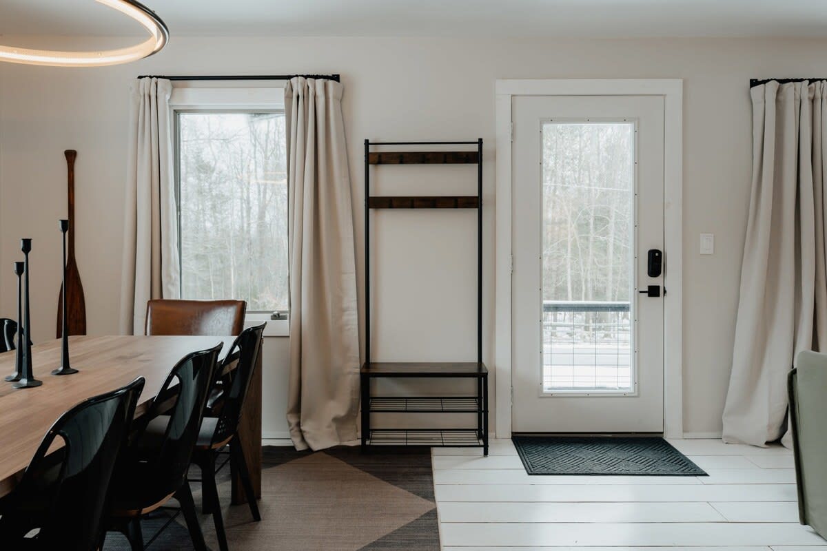 A bright entryway with a white front door featuring a grid window, a dark metal coat rack with shoe storage, and light-colored curtains framing a window looking out to a snowy landscape.