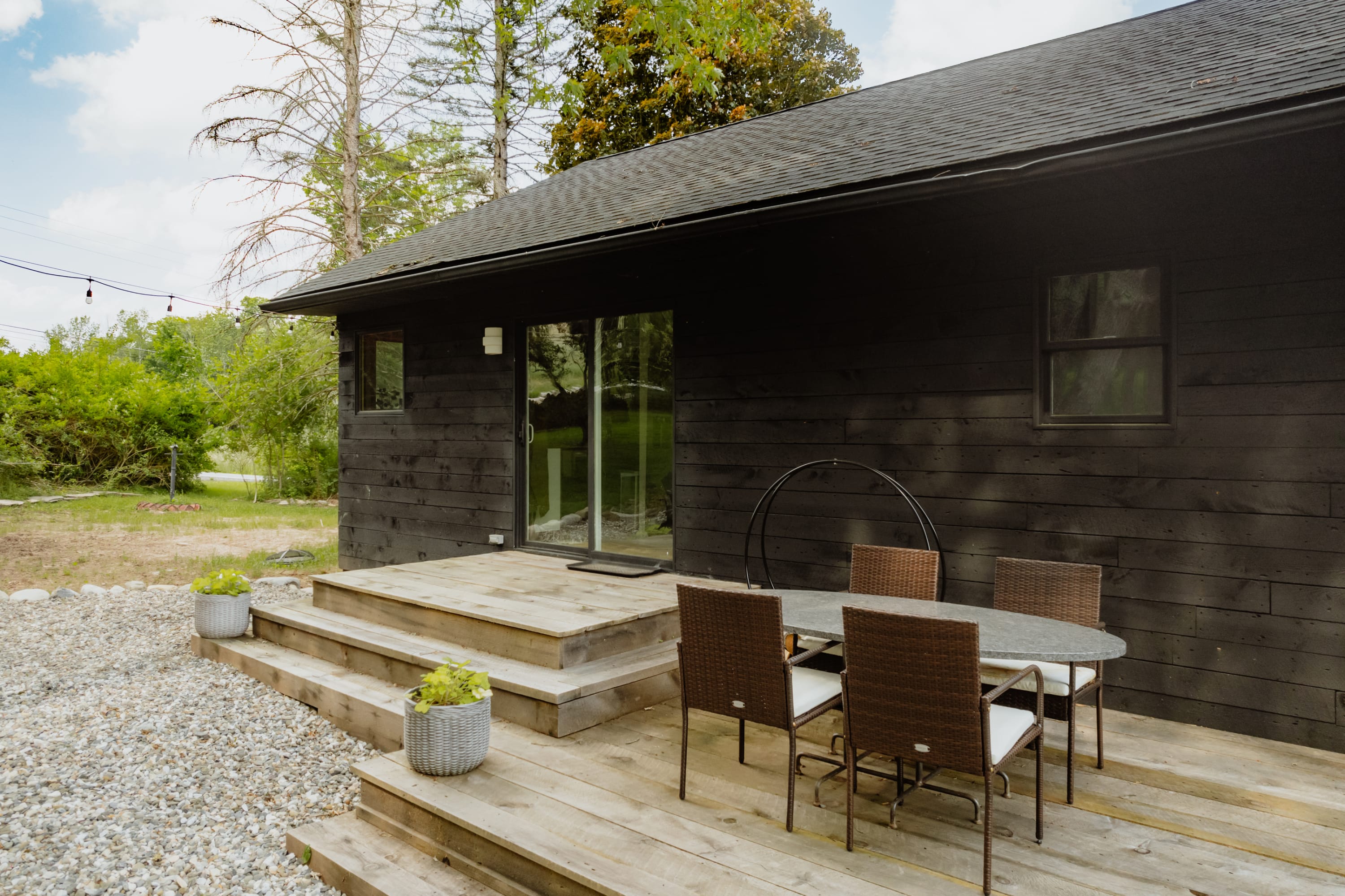 A wooden deck extends from the black-sided cabin, featuring an oval outdoor dining table with four chairs and potted plants. A sliding glass door provides access to the interior.