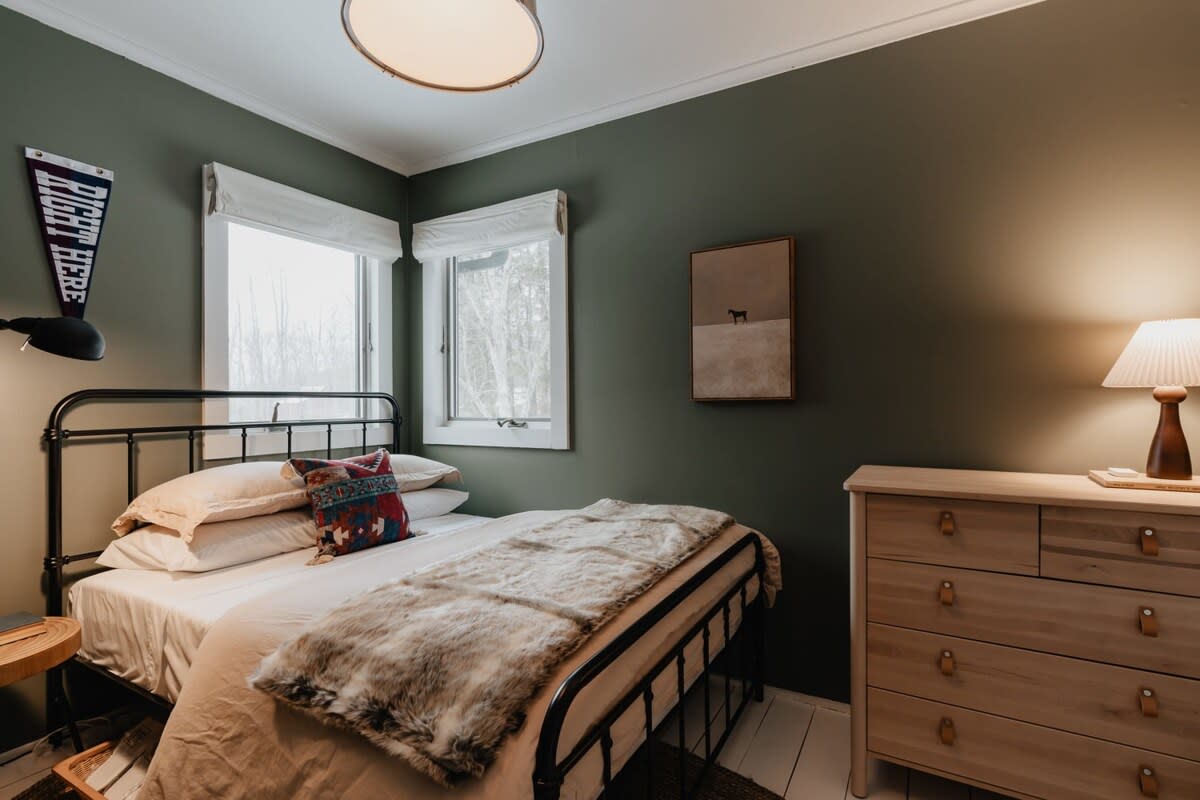 Another view of the cozy bedroom featuring dark green walls, a black metal bed, and a light wooden dresser with a table lamp. Two windows offer natural light, complementing the warm interior.